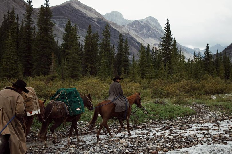 Men on horseback and pack animals ford a rocky stream in a mountain valley, forest in background.