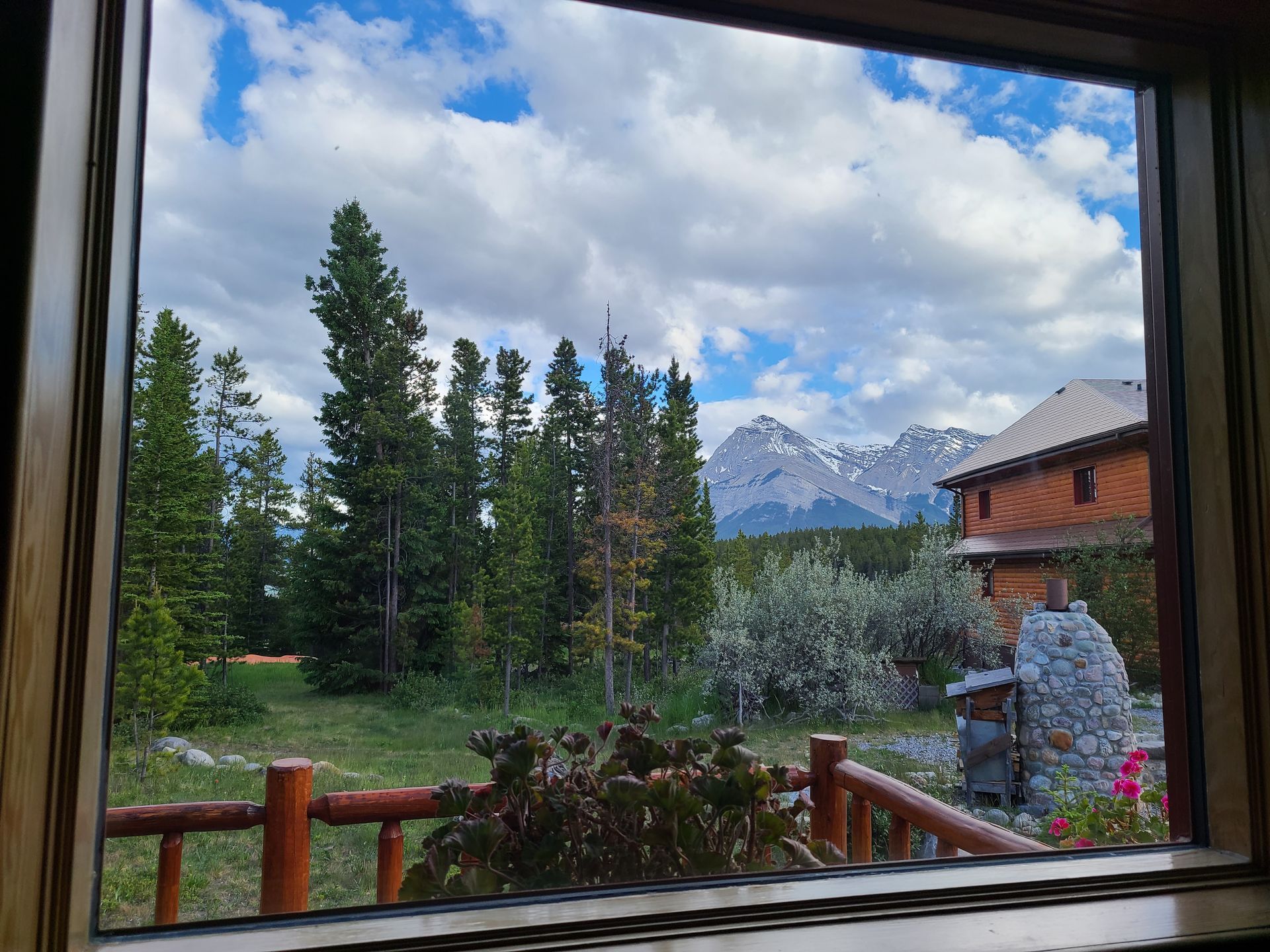View from a window: green trees, grassy yard, wooden lodge, and snow-capped mountains under a cloudy sky.