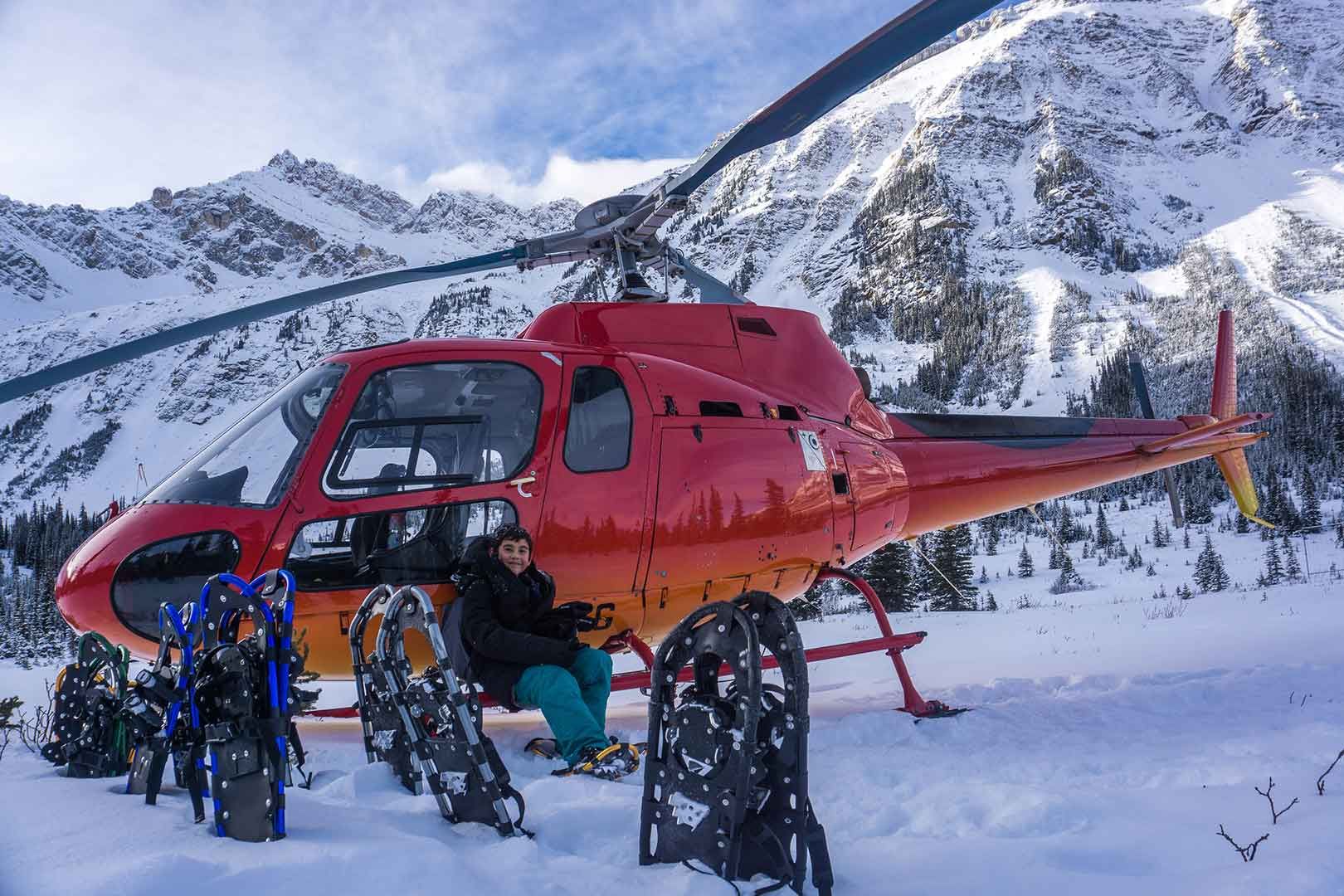 Red helicopter parked in snow, person sits outside. Snowshoes leaned against the helicopter, mountains in background.
