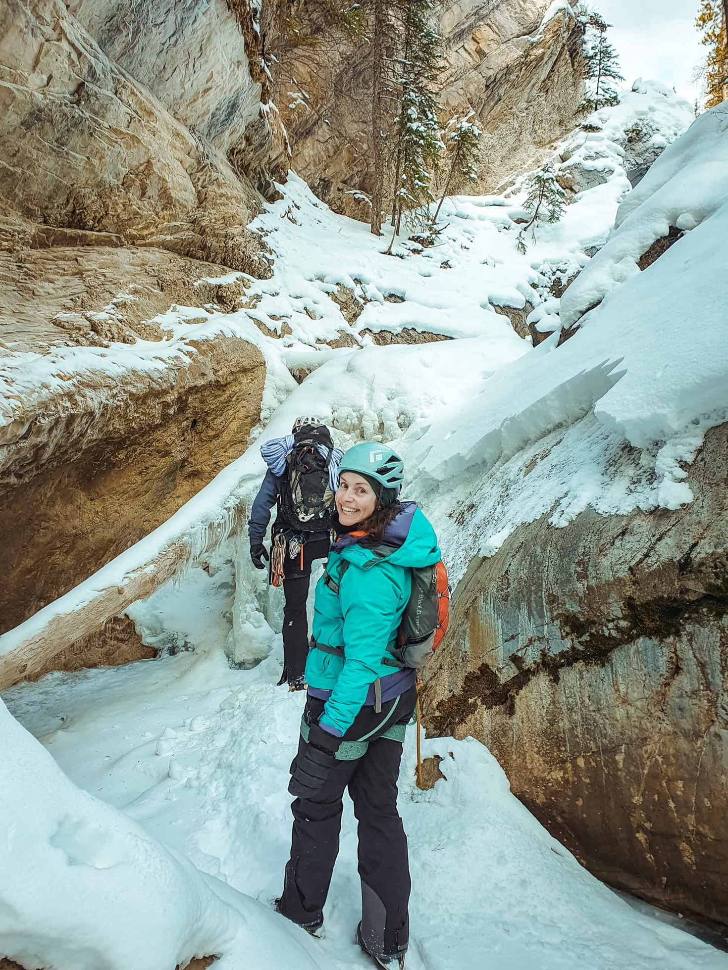 Two hikers in winter gear, walking through a snowy canyon. One smiles at the camera.