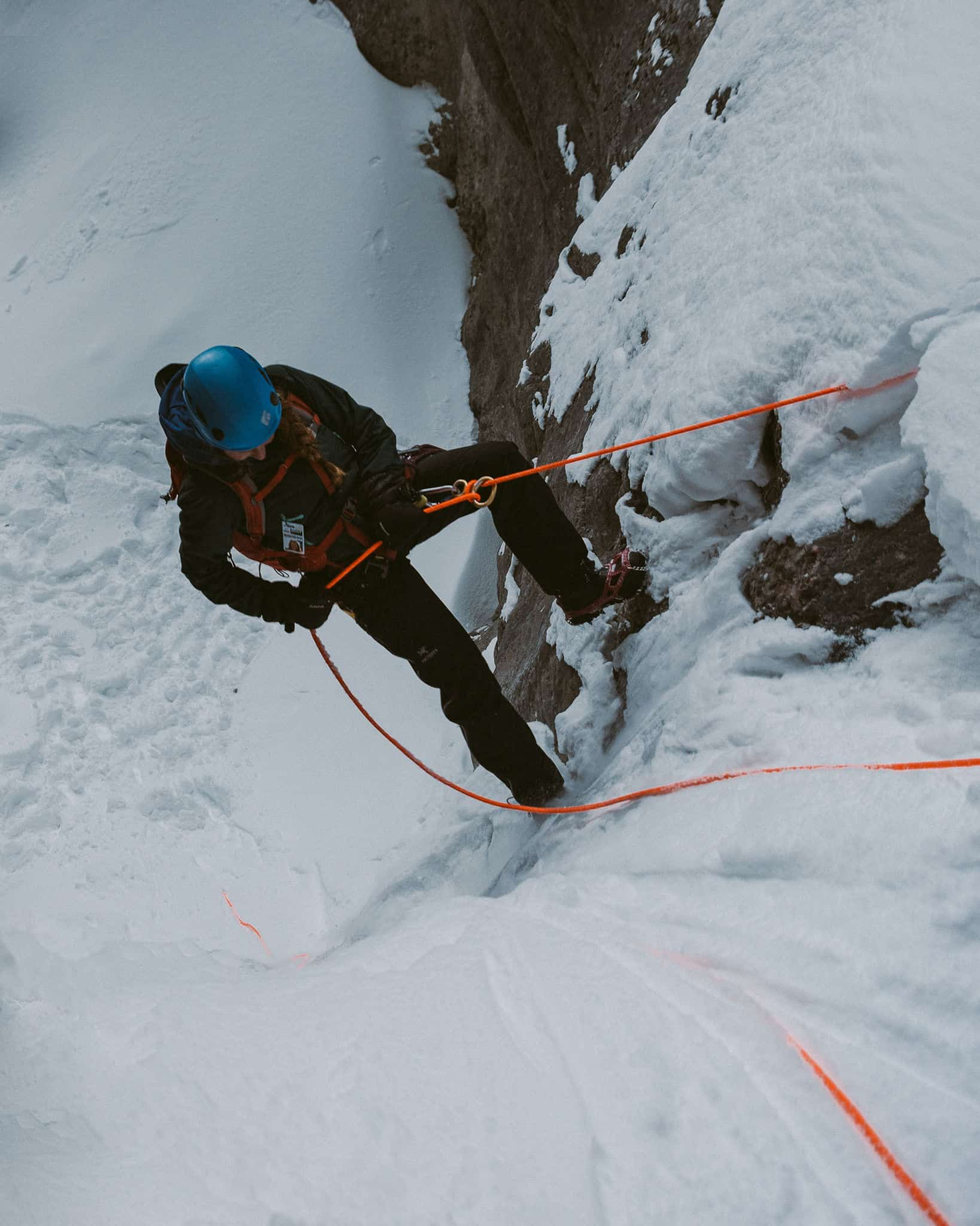 Person rappelling down a snowy cliff face, secured by orange ropes, wearing a blue helmet and dark clothing.