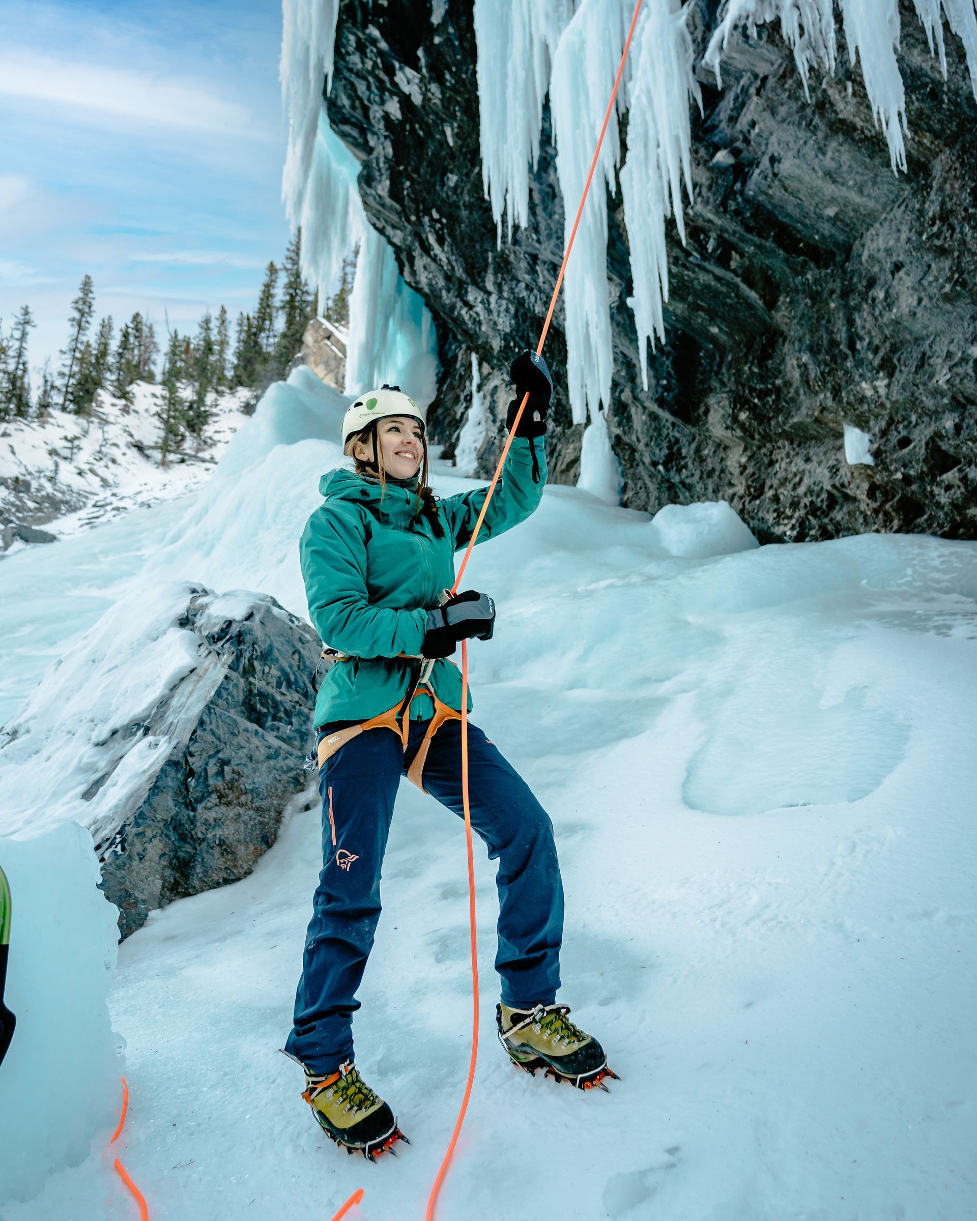 Woman ice climbing, turquoise jacket, blue pants, helmet, orange rope, icy cliff, snowy ground.