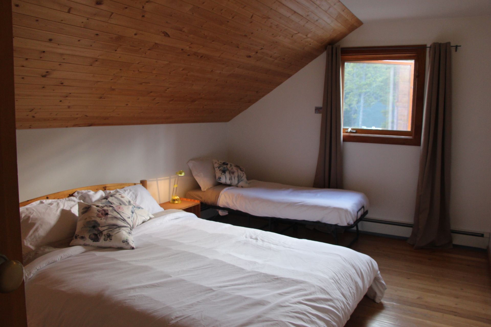 Bedroom with two beds under a sloped, wood-paneled ceiling; one window with curtains.