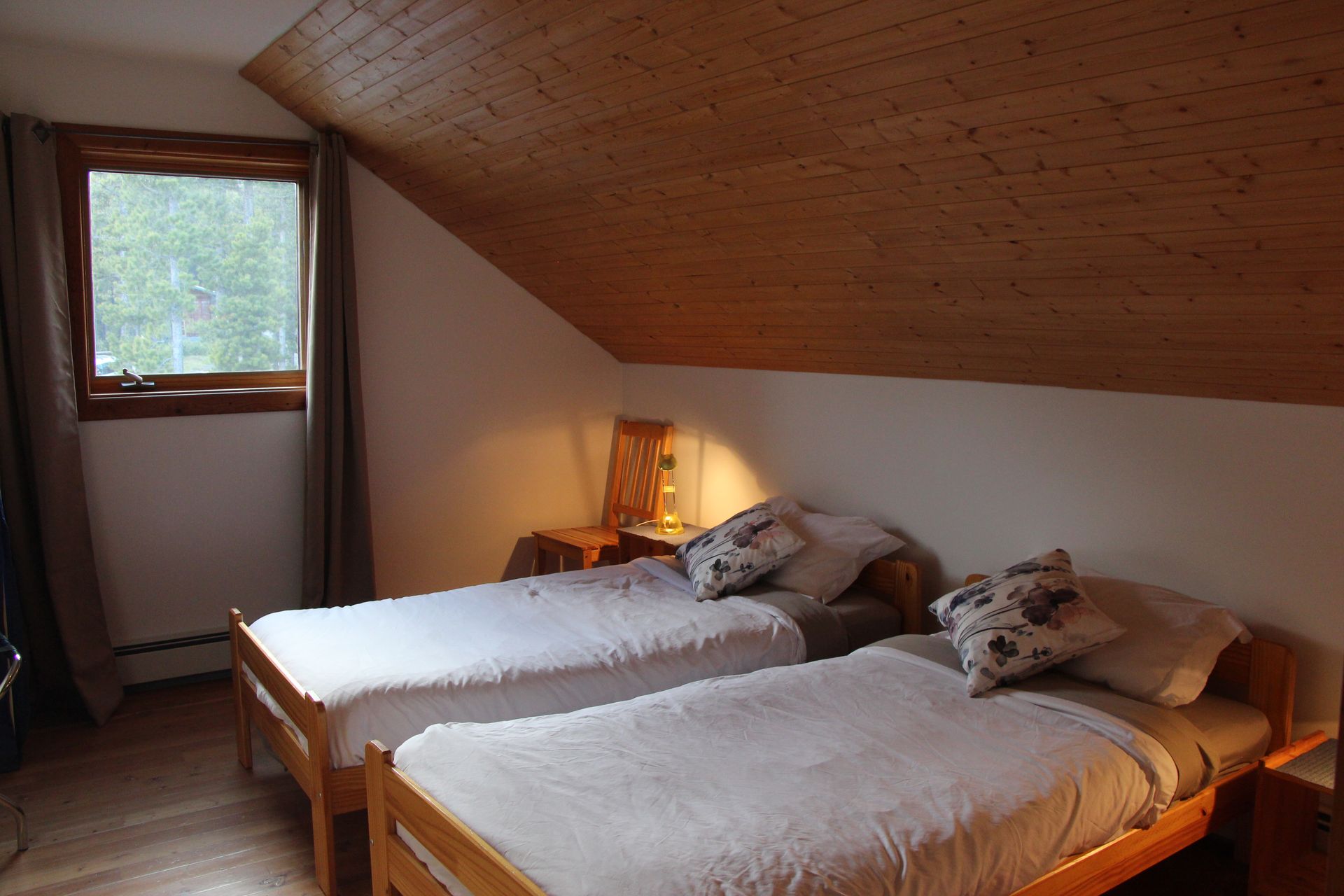 Two twin beds in a room with a sloped wooden ceiling. A window with curtains is visible.