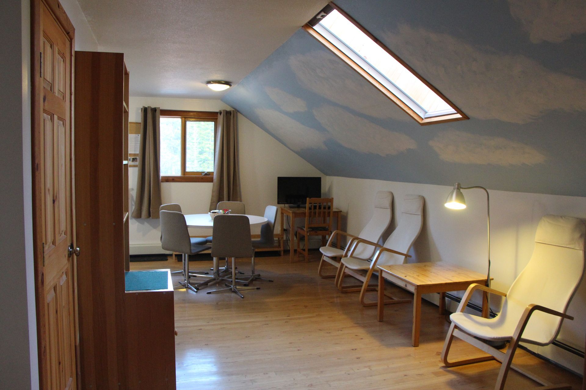 Attic room with wood floor, sky-painted ceiling, chairs, table, small tv, and a skylight.