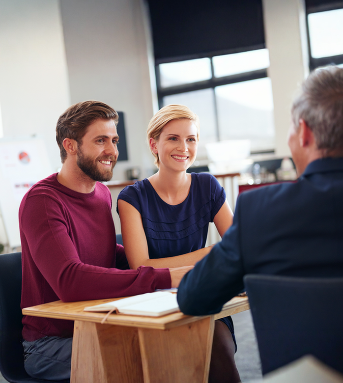 A man and a woman are sitting at a table talking to a man in a suit.