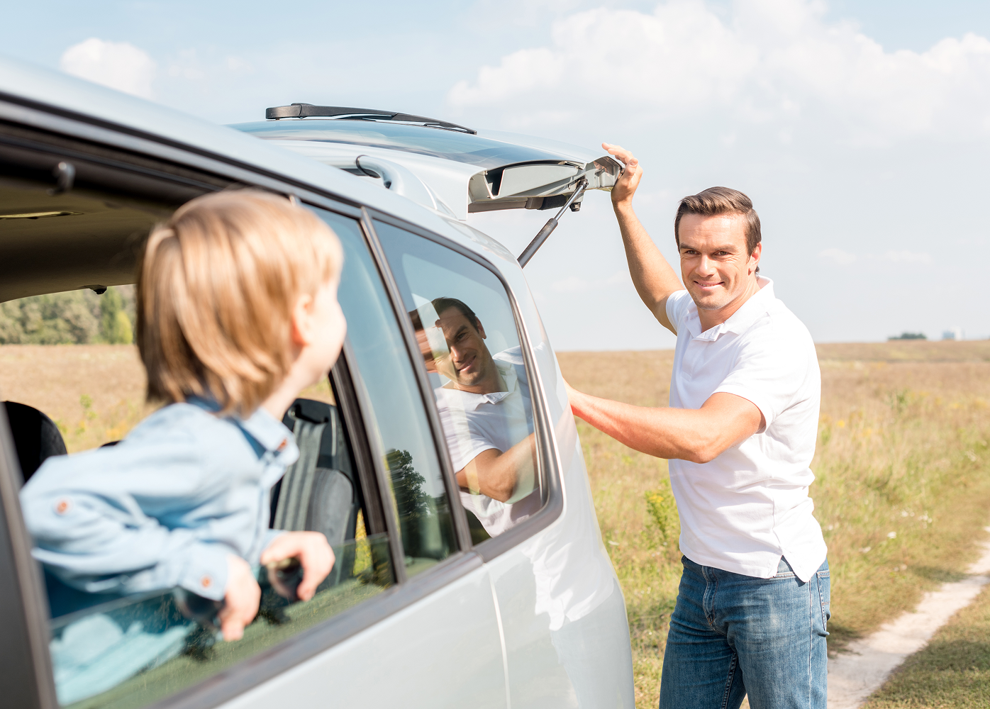 A man is opening the trunk of a car with a child looking out the window.