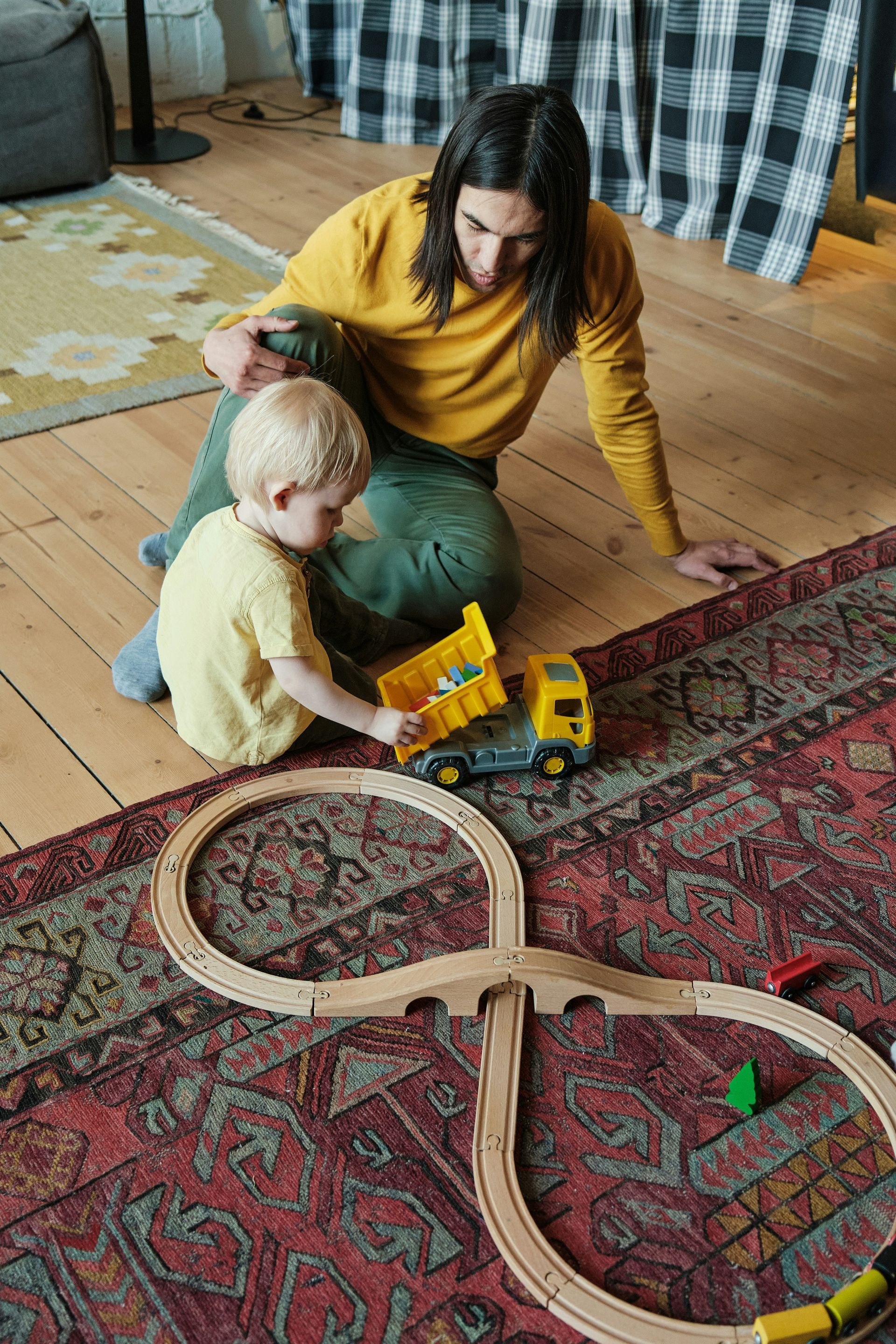 A person in a yellow sweater and a toddler play with a toy dump truck and a wooden track on a red patterned rug.