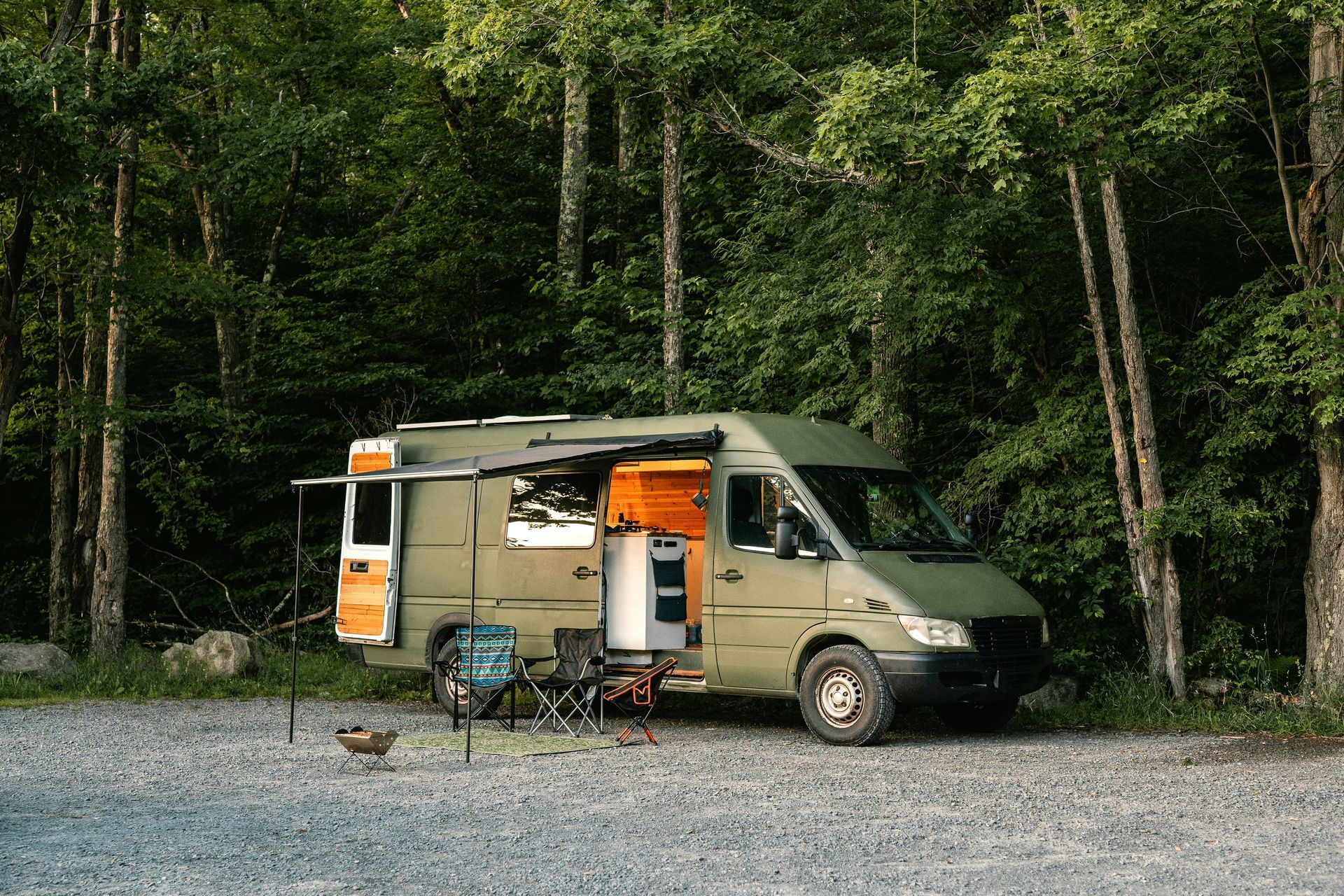 A matte green camper van parked in a forest clearing with its side door open, an awning extended, and outdoor furniture.