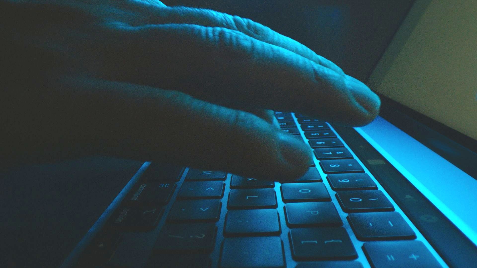 A hand typing on a laptop keyboard in low light, illuminated by a cool blue glow.