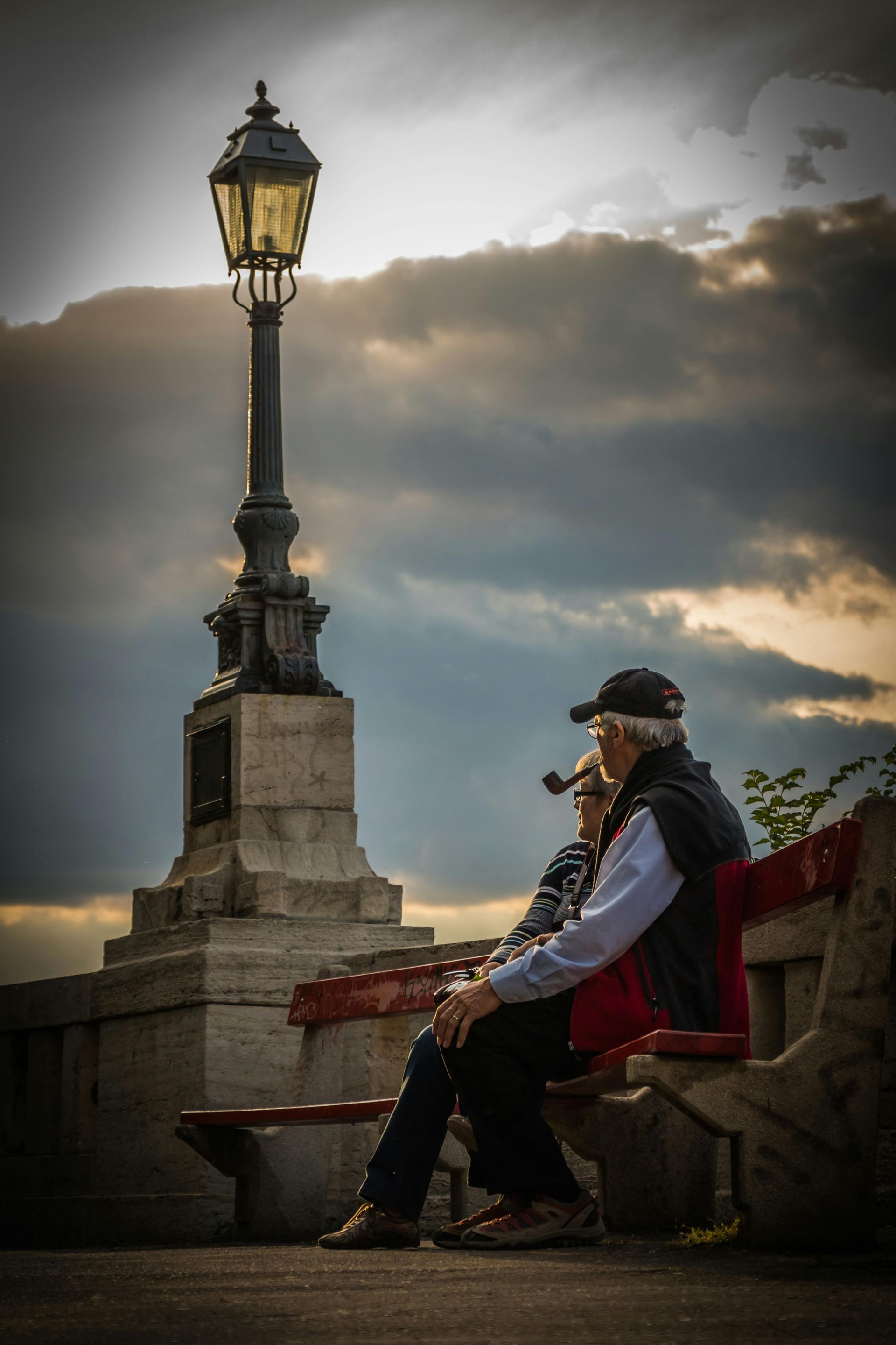 An older person with a pipe sits on a stone bench beside an ornate lamppost under a dramatic, cloudy sunset sky.