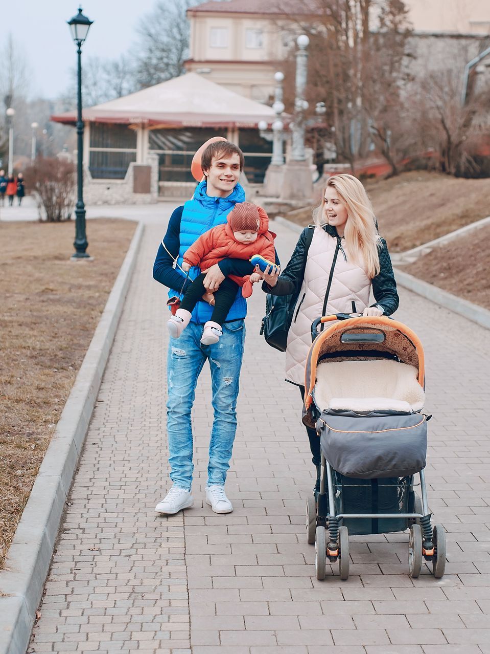 A family walks on a park path, with one person carrying a child and the other pushing a stroller.