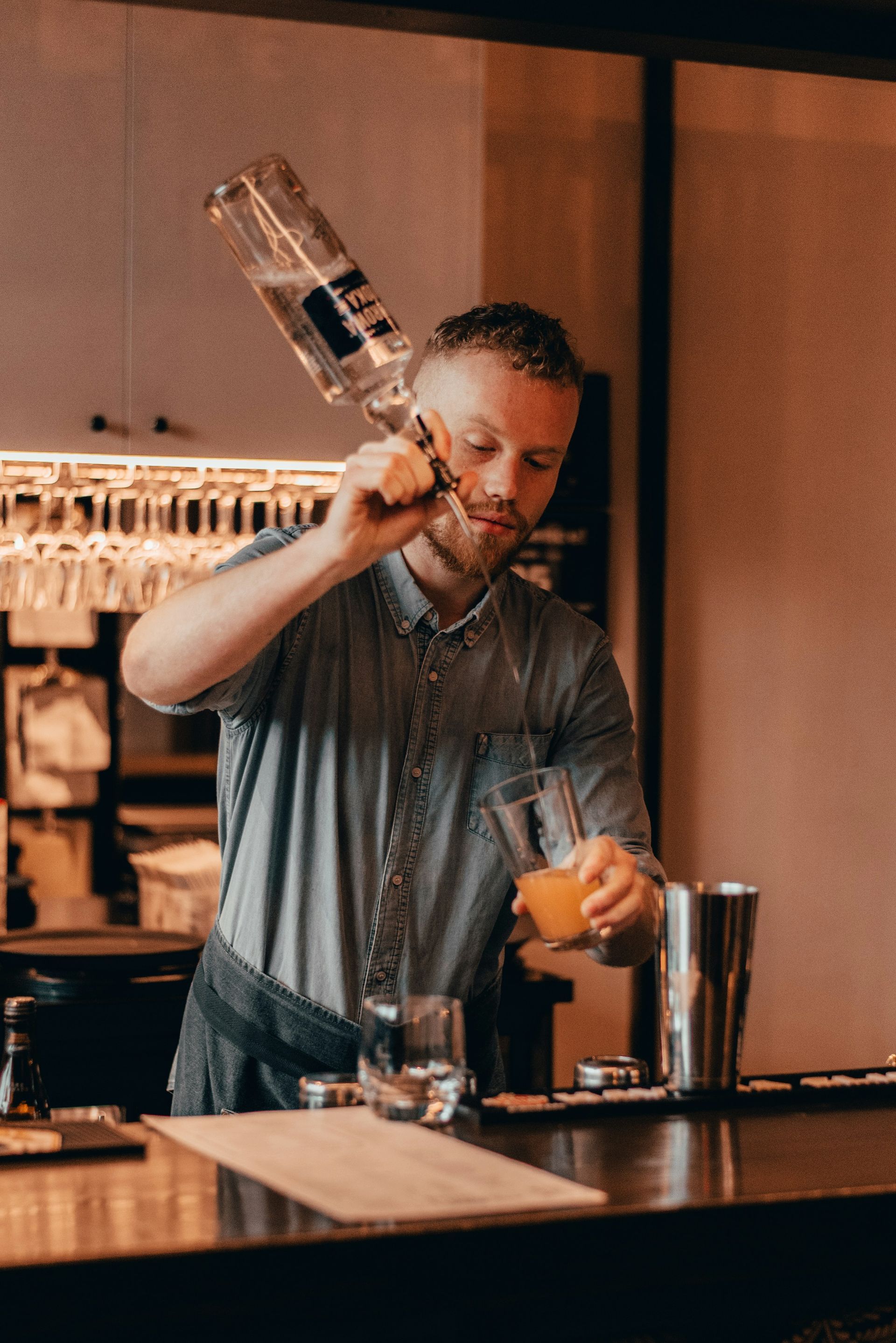 A bartender in a gray shirt pours liquid from a bottle into a glass at a wooden bar counter.