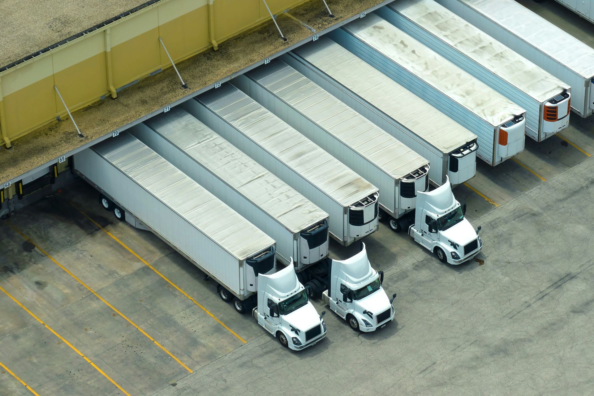 An aerial view of several white semi-trucks parked in a row at a warehouse loading dock.
