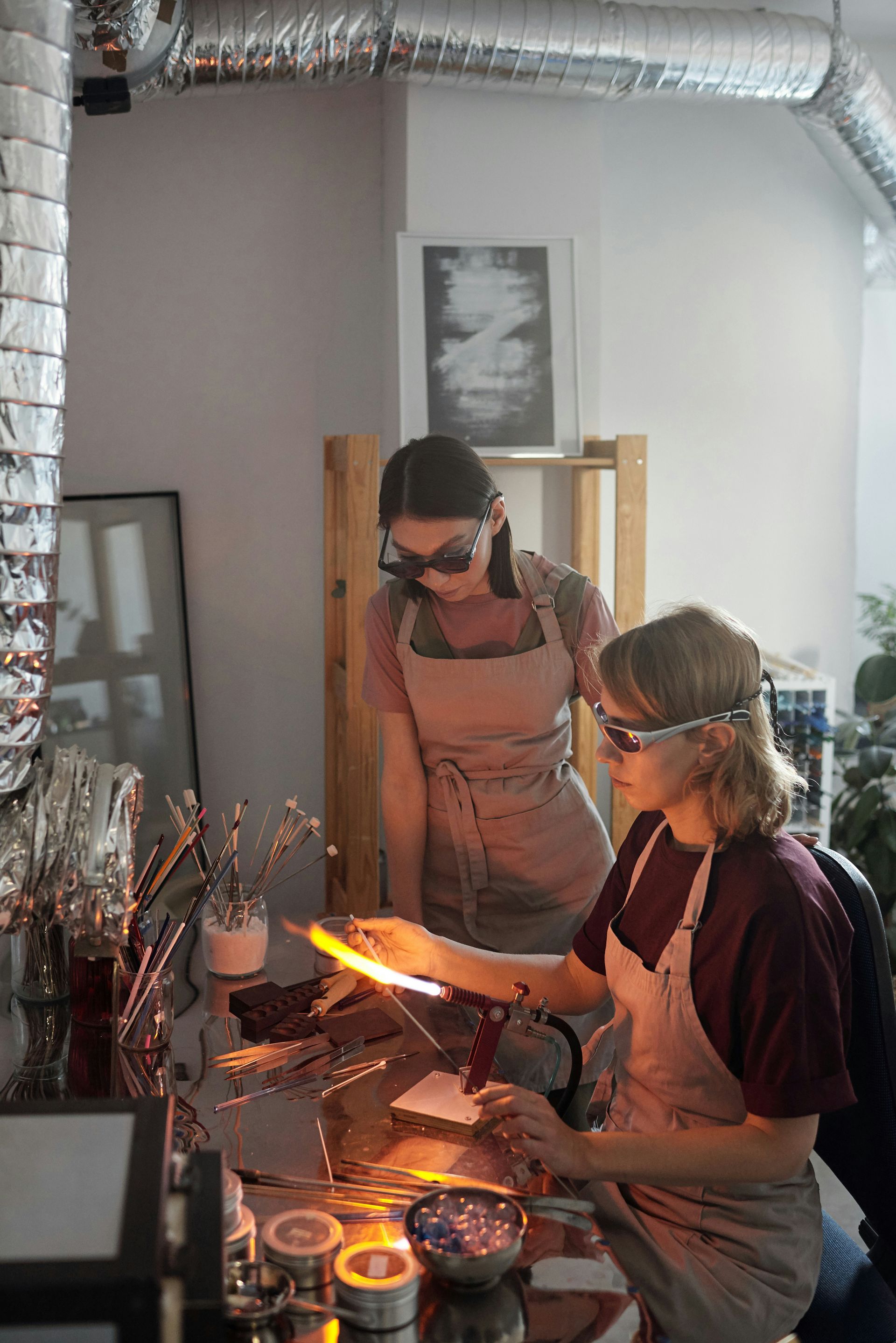 Two people wearing protective eyewear and aprons work with a flaming torch at a glassworking studio.