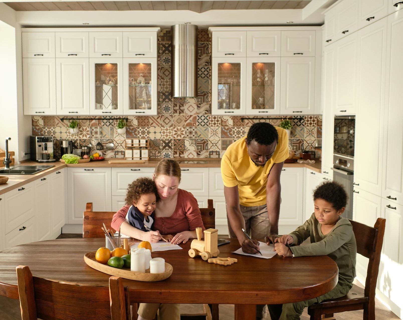 A family sits at a wooden kitchen table, working on papers together in a bright, modern, white-cabinet kitchen.