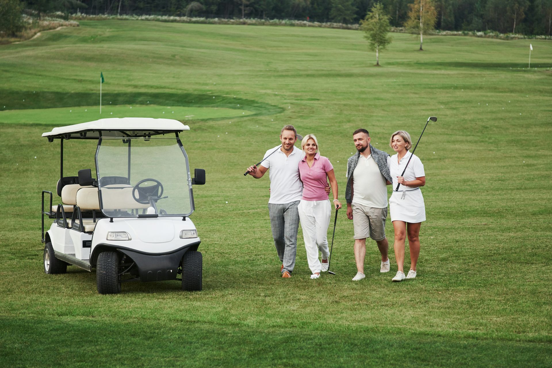 Four people walk and talk on a golf course near a parked white golf cart, holding clubs and smiling.