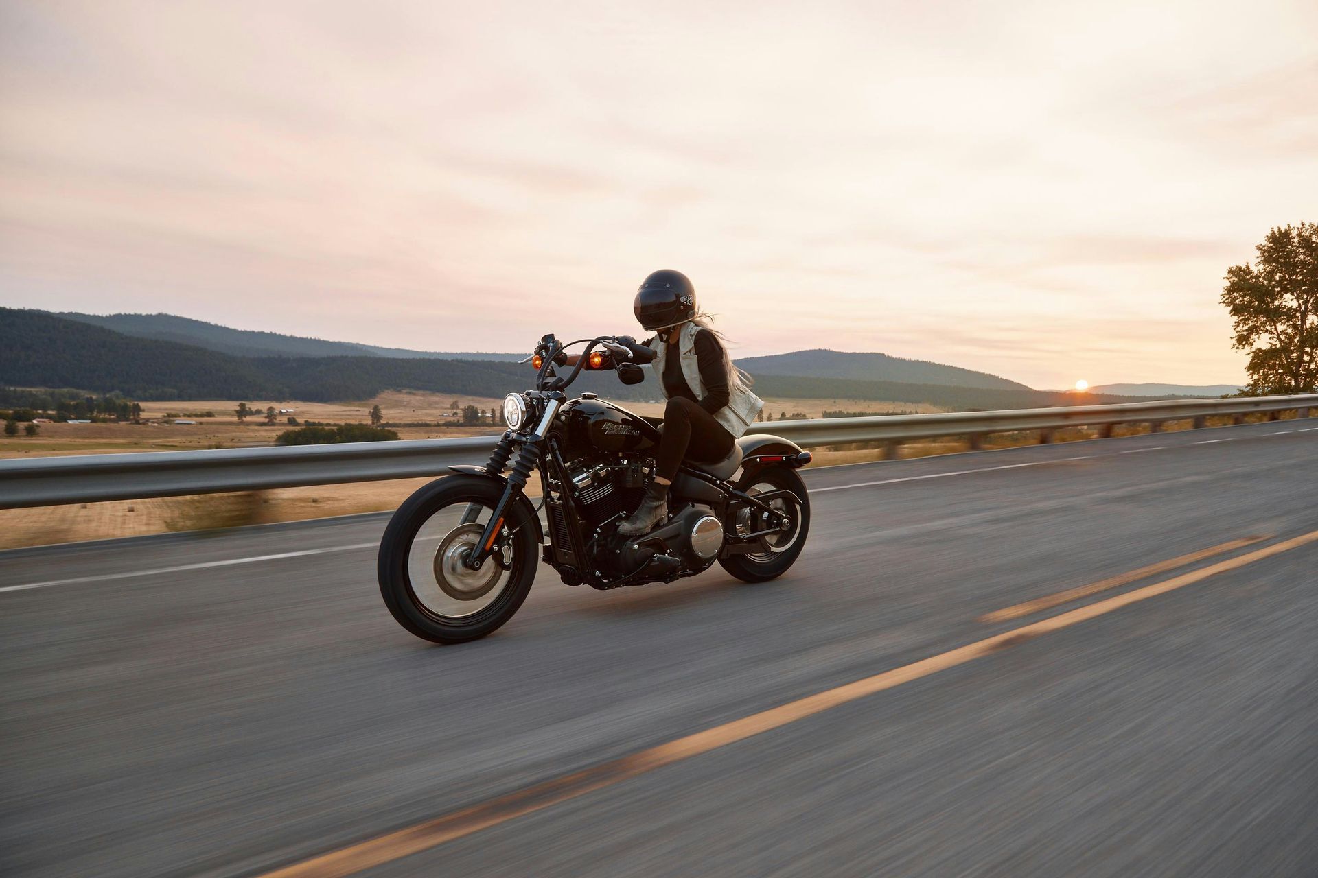 A person in a black helmet rides a black cruiser motorcycle on a highway during a scenic sunset.
