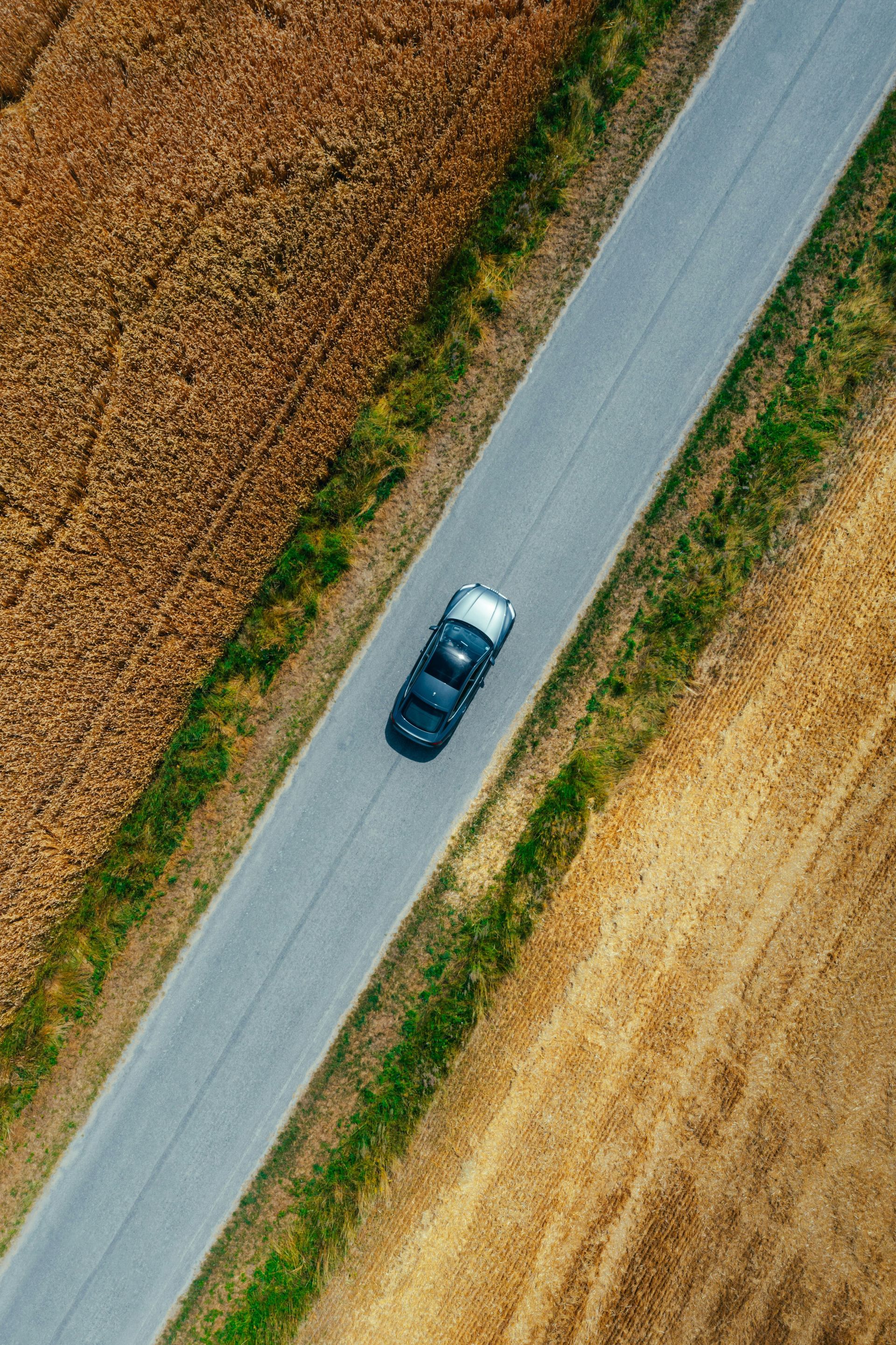 An aerial view of a dark blue car driving on a straight, paved road through golden, harvested fields.
