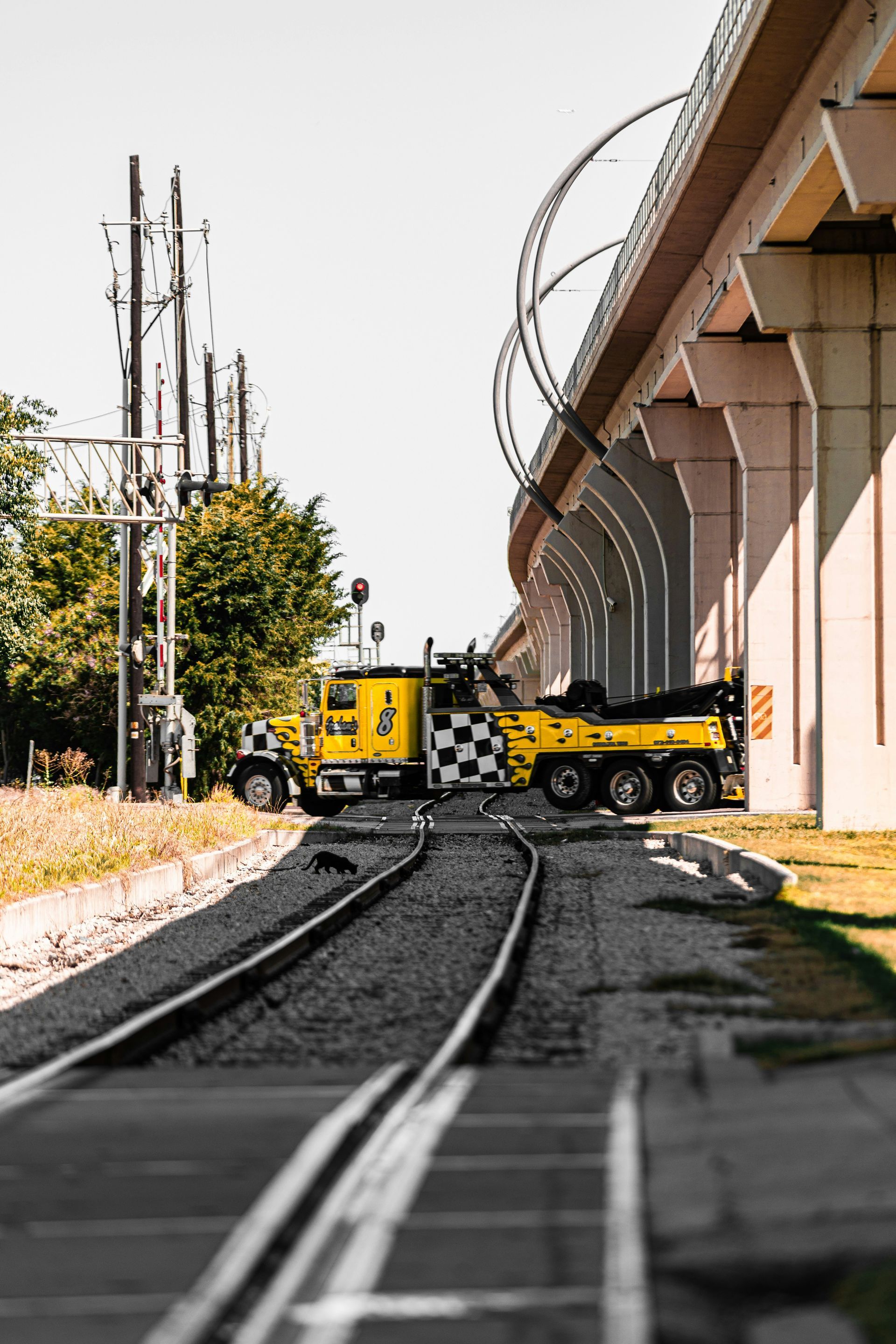 A yellow tow truck parked on train tracks beneath a highway overpass.