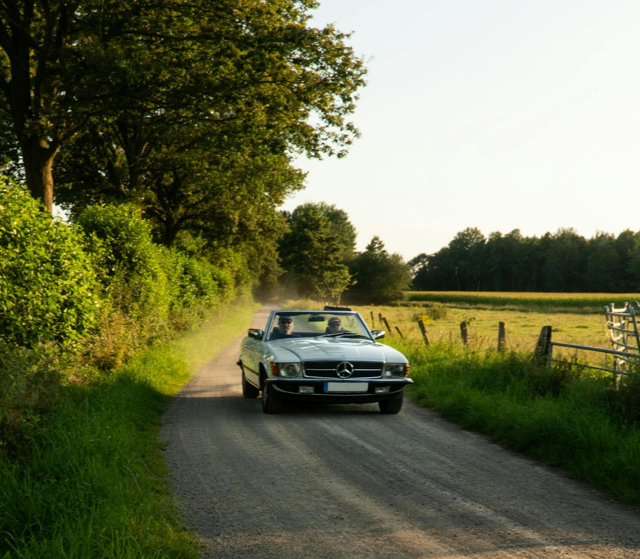 A silver vintage convertible car drives down a narrow, sunlit country road lined with green trees and fields.