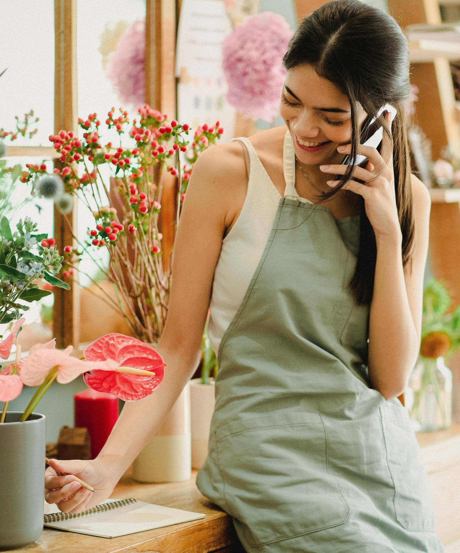 A person wearing a sage green apron smiles while talking on a phone, standing in a flower shop near a notebook.