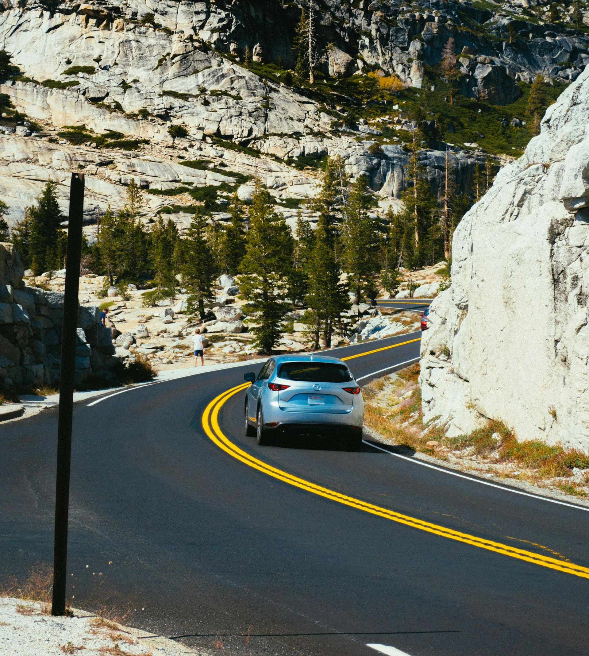 A silver car drives along a curved mountain road flanked by rocky cliffs and pine trees under bright sunlight.