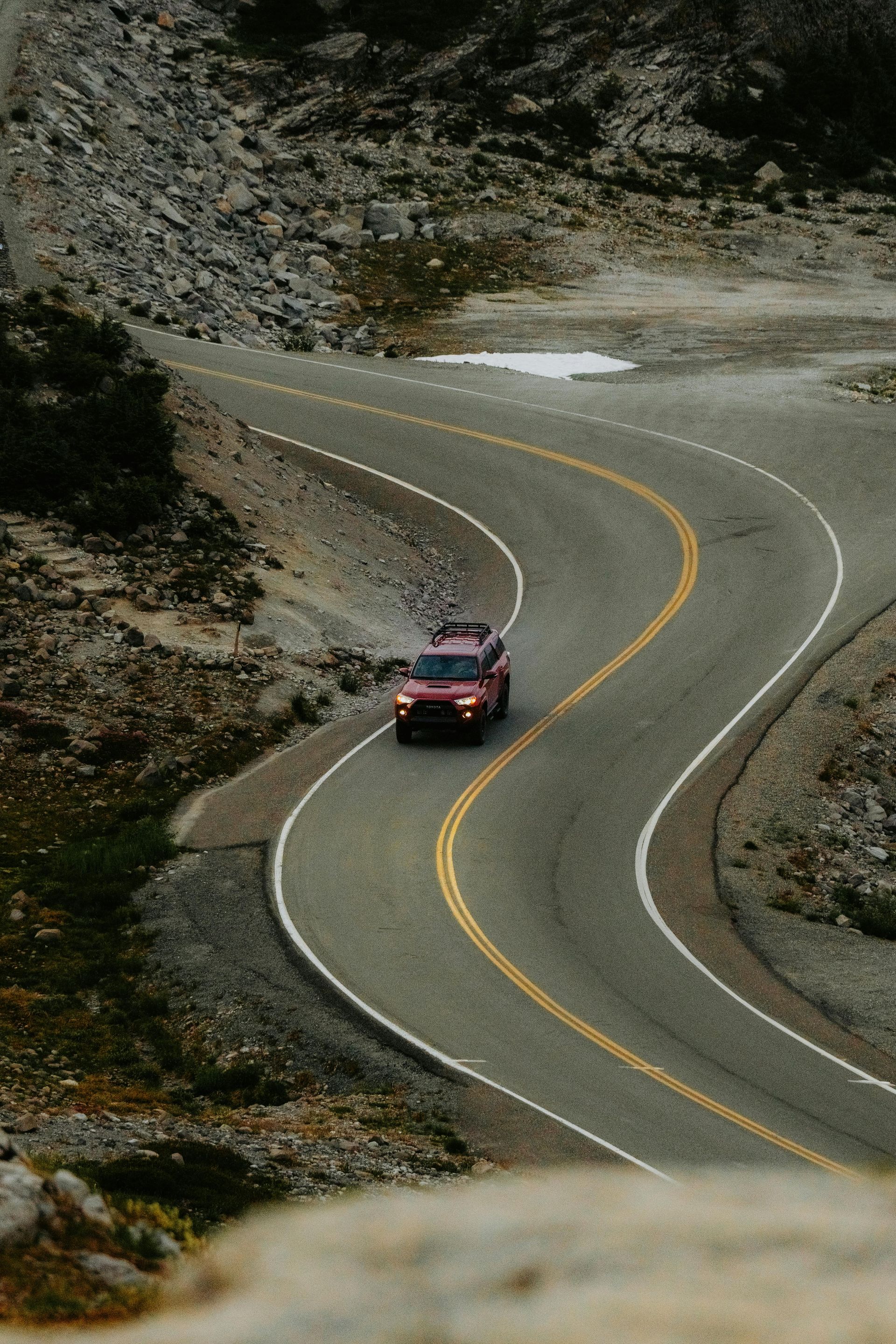 A red SUV drives along a curving, paved mountain road surrounded by rocky, barren terrain.