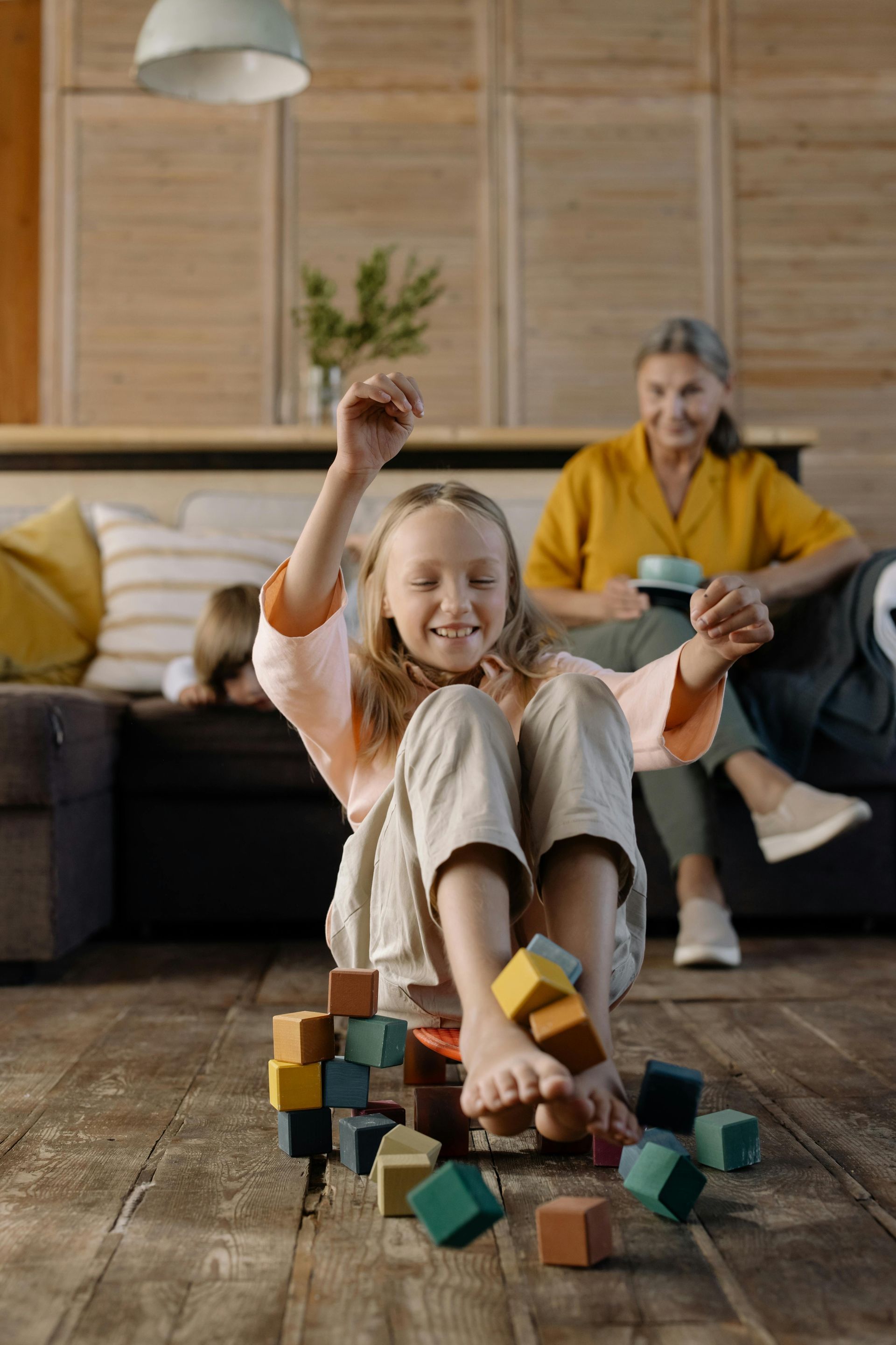 A joyful child sits on a wooden floor playing with colorful building blocks, with an adult watching from a couch nearby.