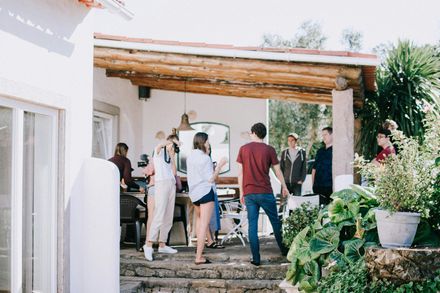 A group of people gathers on a sunny stone patio under a wooden pergola attached to a white house.