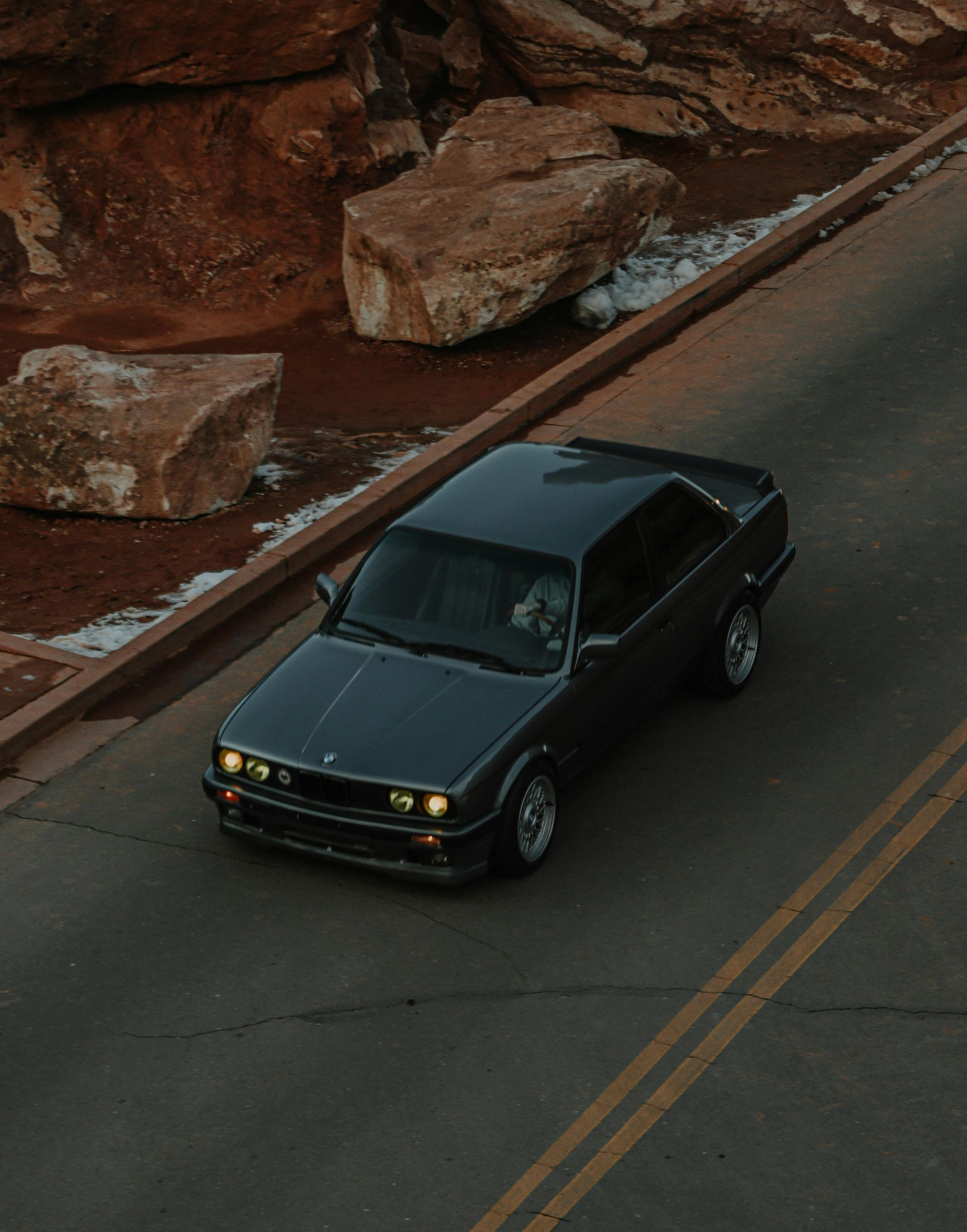 A dark gray vintage BMW sedan drives along an asphalt road next to large, rocky red cliffs.