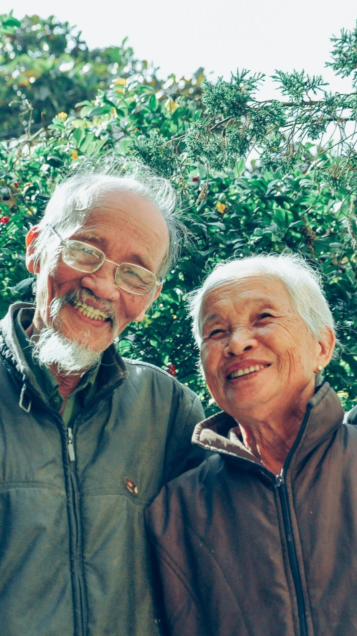 Two smiling people wearing zip-up jackets stand together outdoors in front of a lush green tree.