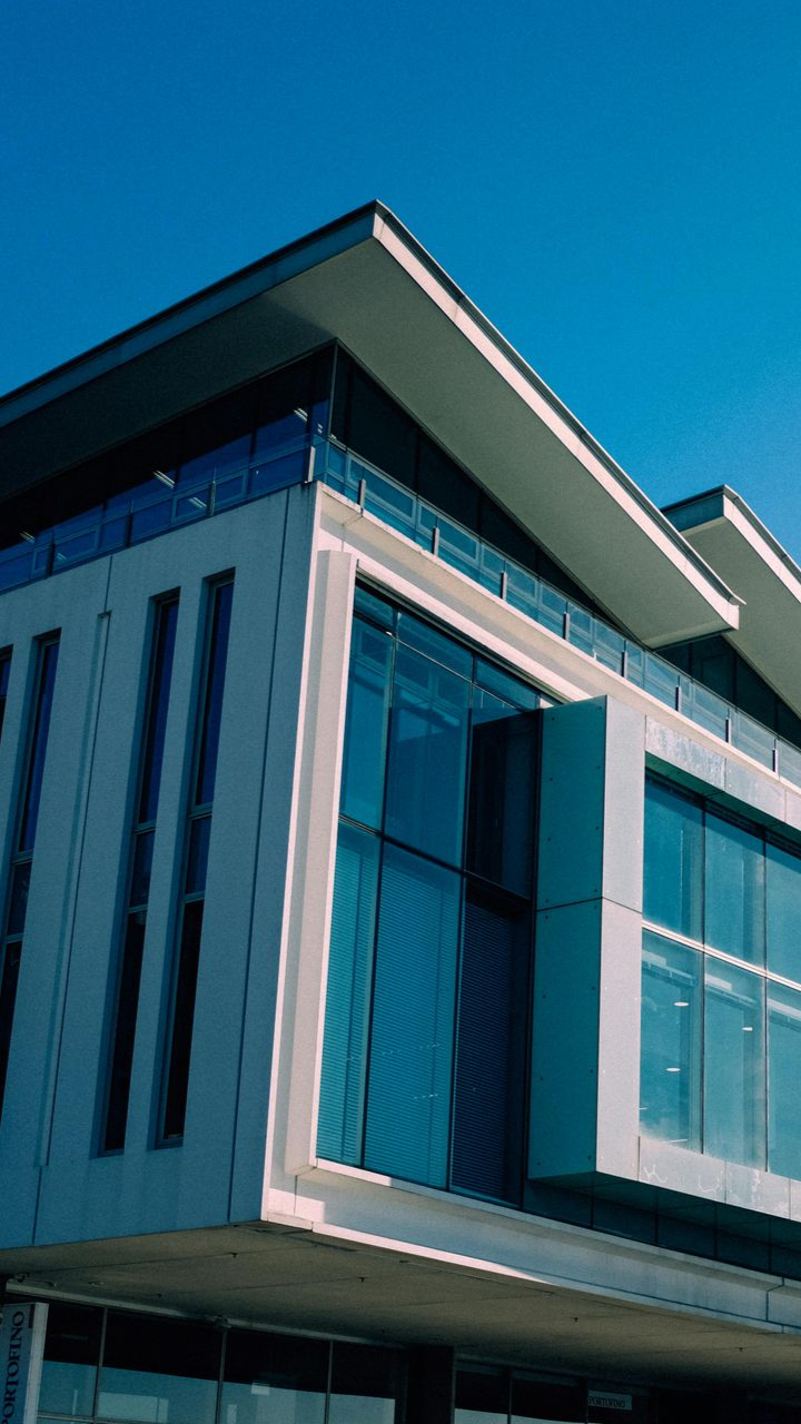 A modern multi-story building featuring large glass windows, white paneling, and an angled roof under a clear blue sky.