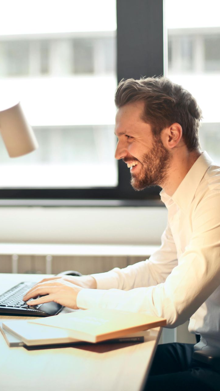 A smiling professional in a white shirt types on a keyboard at a desk in a sunlit office.