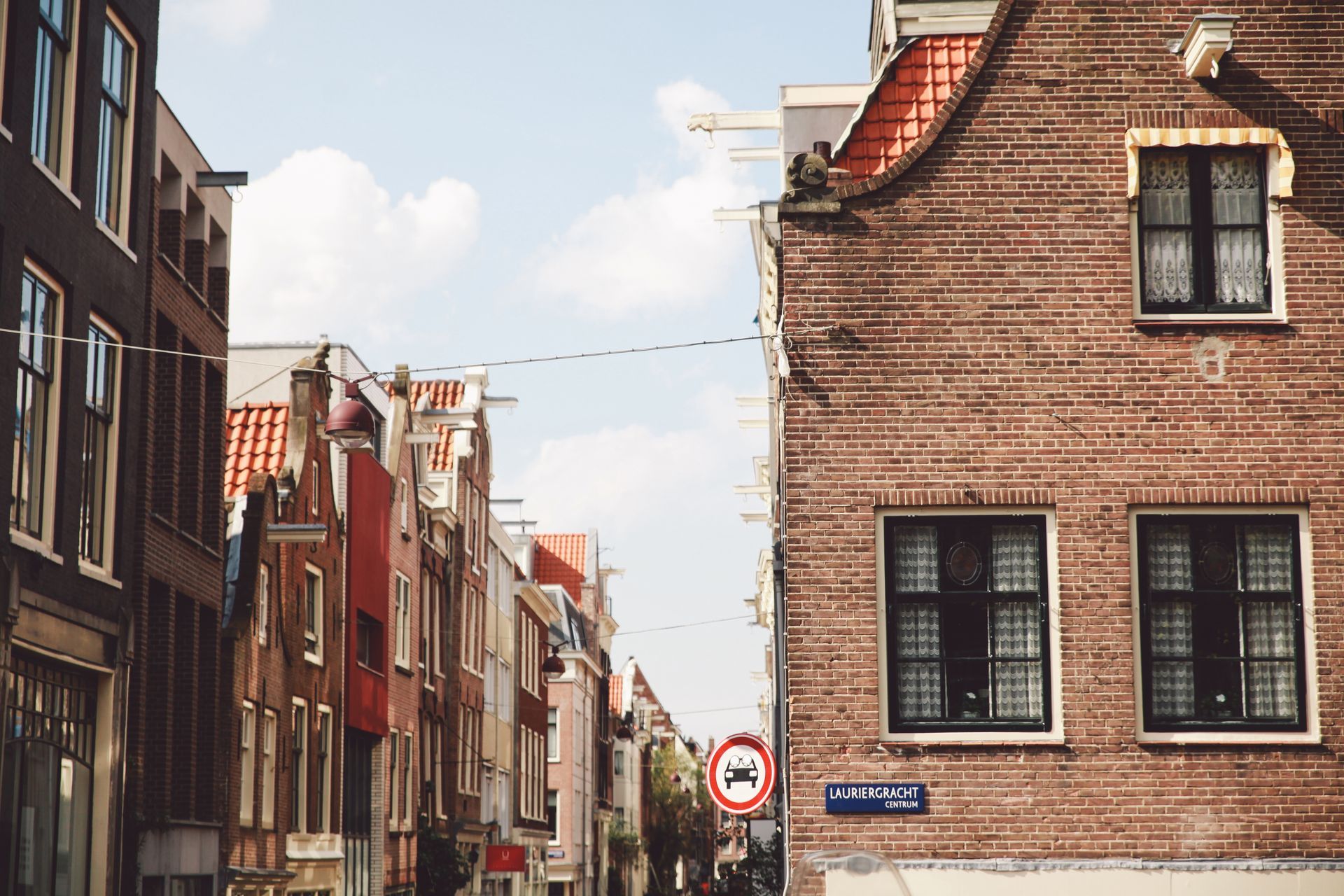 A street view in Amsterdam with historic narrow brick buildings on both sides, a red circular traffic sign, and blue sky.