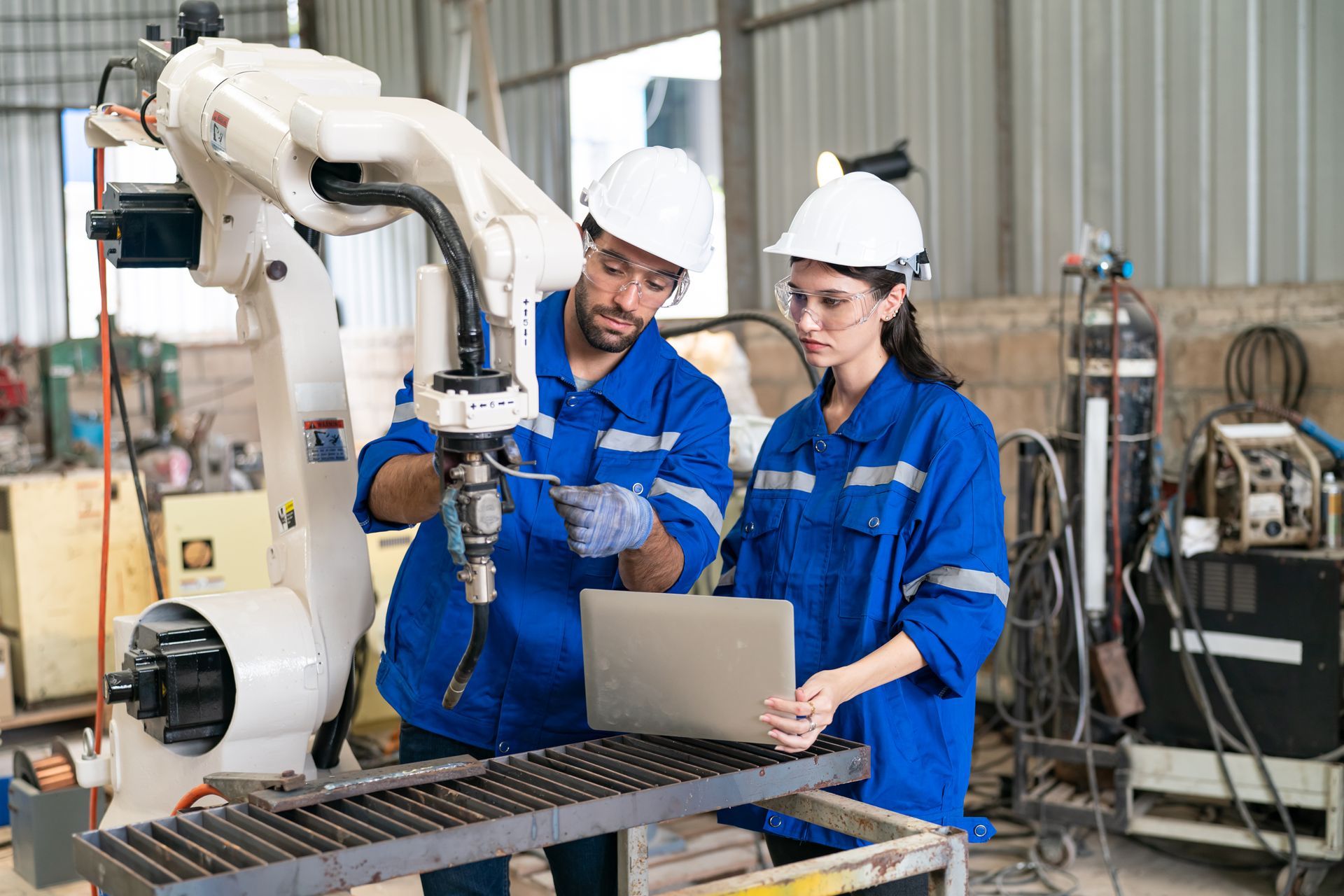 Two technicians in blue uniforms and hard hats inspect a robotic arm while consulting a laptop in an industrial workspace.