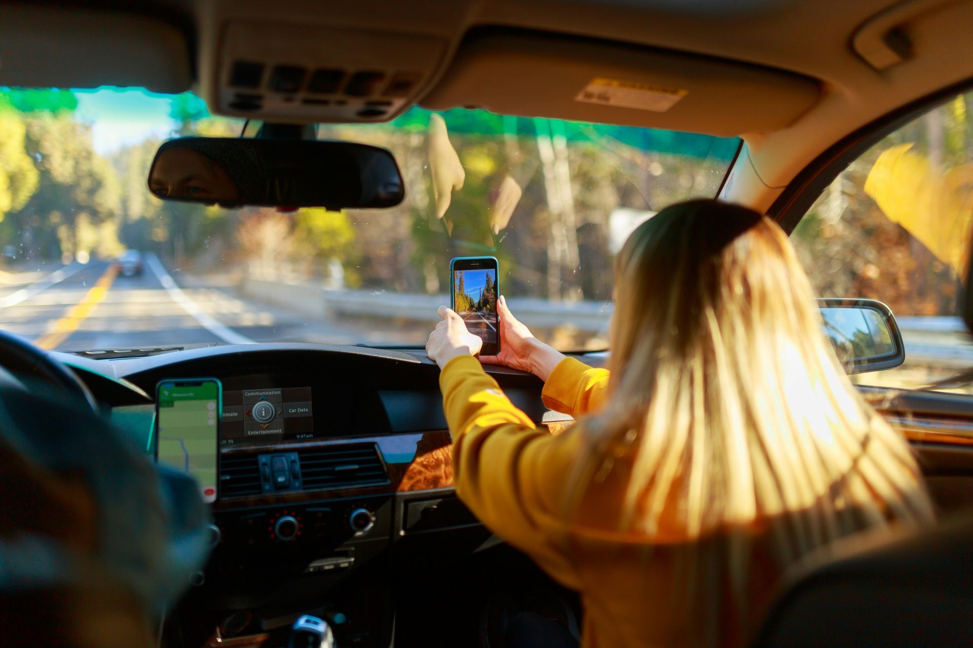 A person in a yellow sweater sits in a car, holding up a smartphone to photograph the scenic road ahead.
