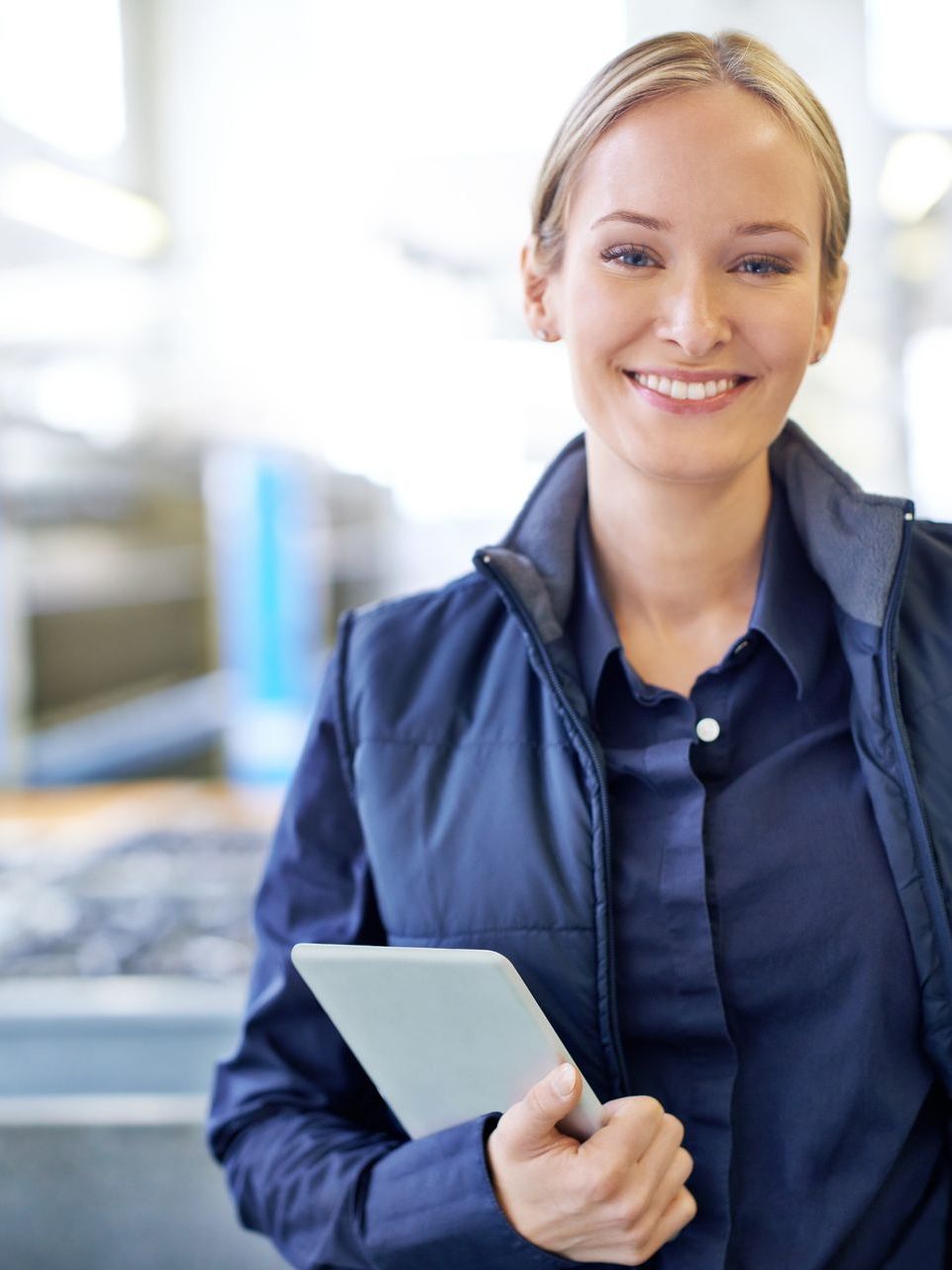 A smiling professional in a blue collared shirt and vest stands in a workplace, holding a tablet.