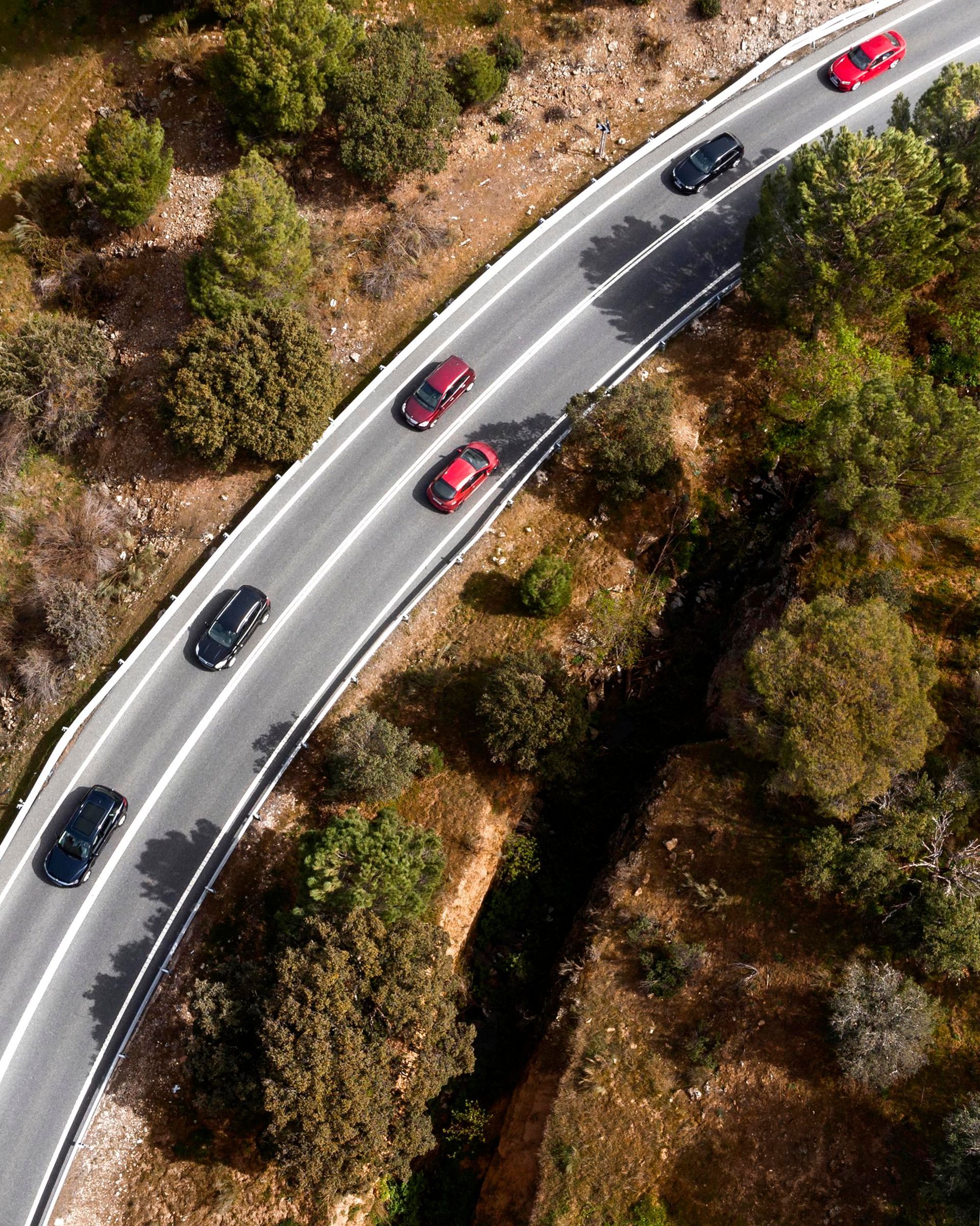 An aerial view of cars in red and black driving along a winding asphalt road through a wooded landscape.