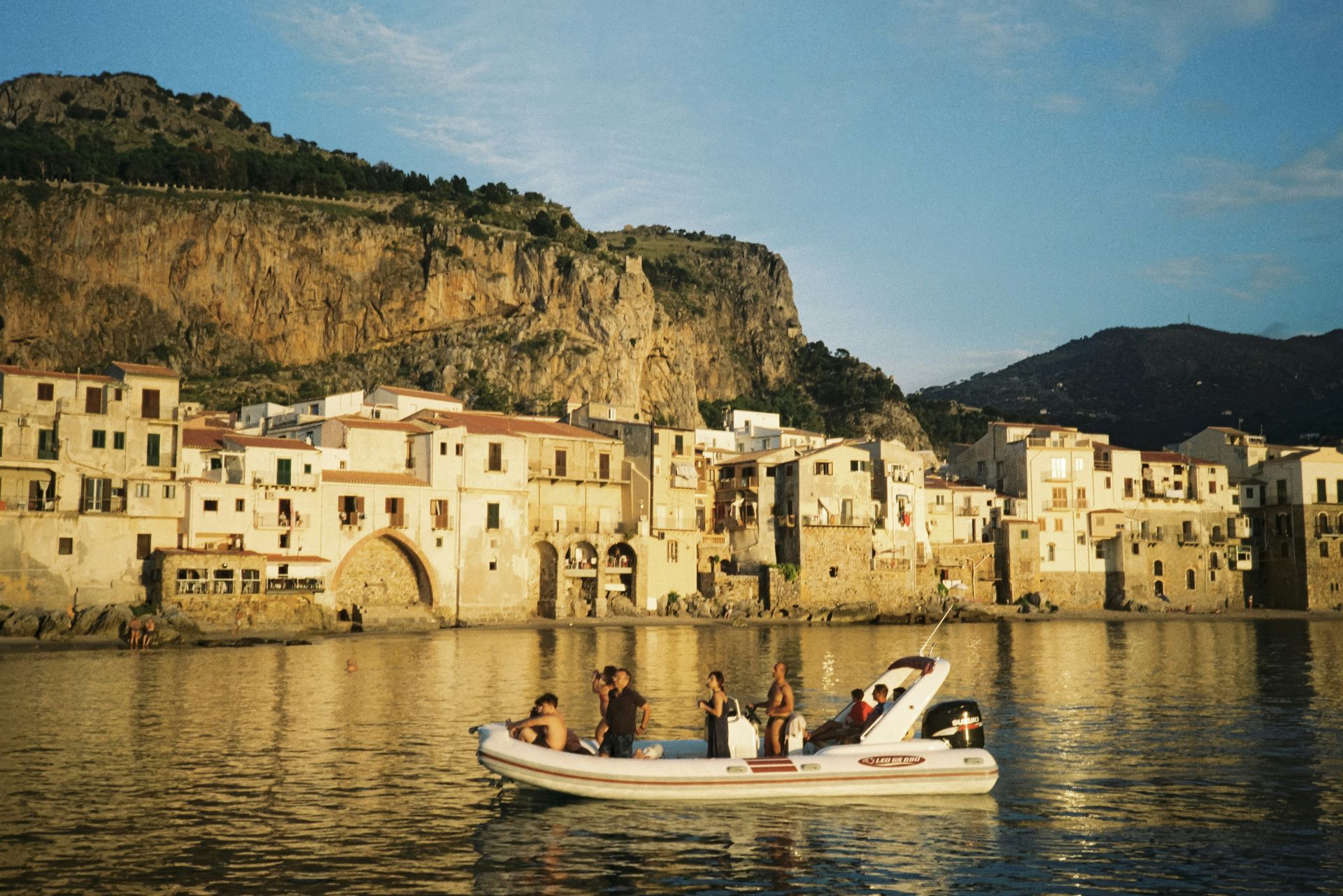A rigid inflatable boat with passengers near the coastline of Cefalù, Sicily, with historic buildings below a mountain.