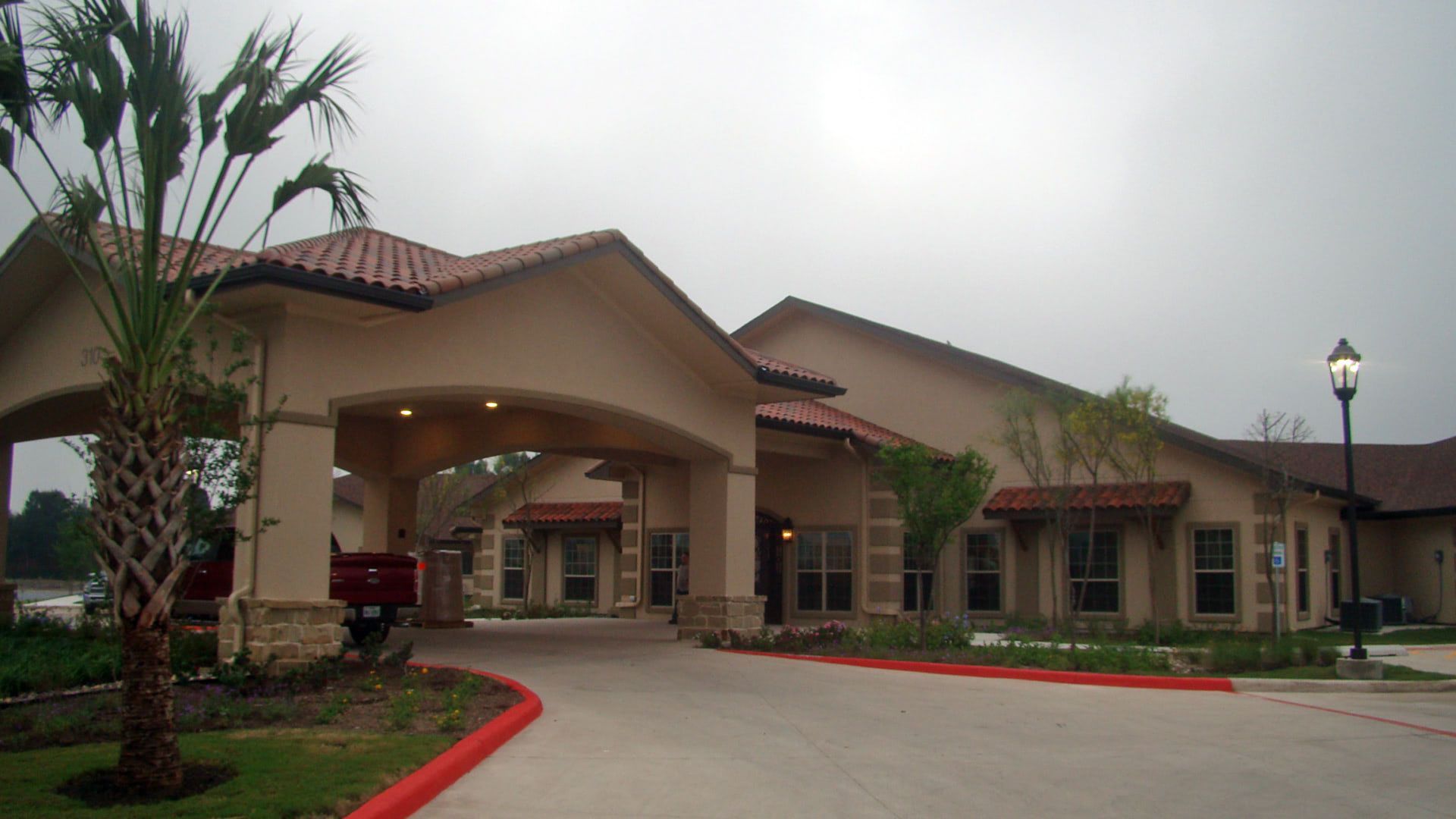 Covered entrance of a beige building with red tile roof and landscaped driveway under cloudy sky