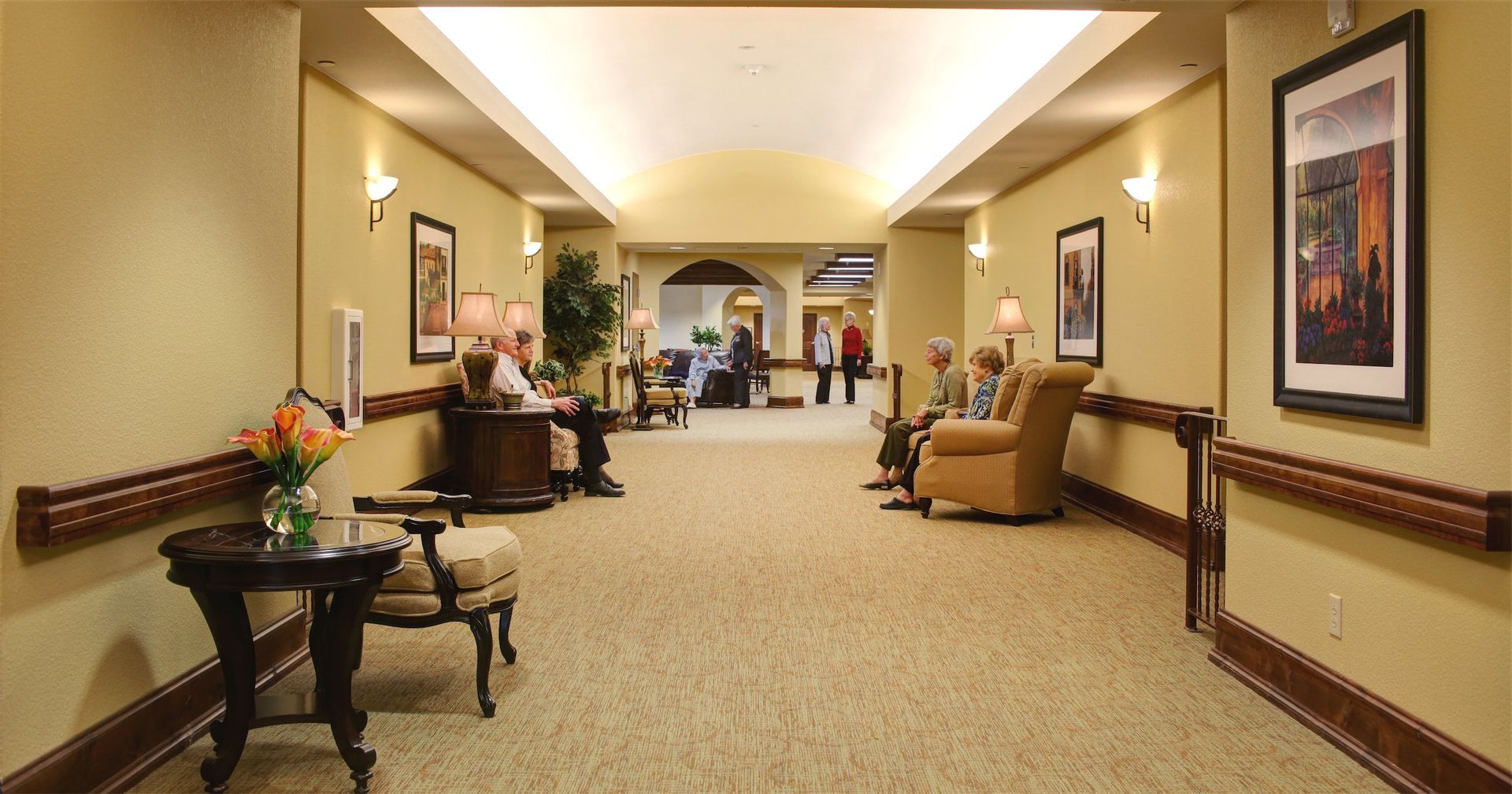 Hallway in a care facility with residents sitting in chairs and a table with flowers.