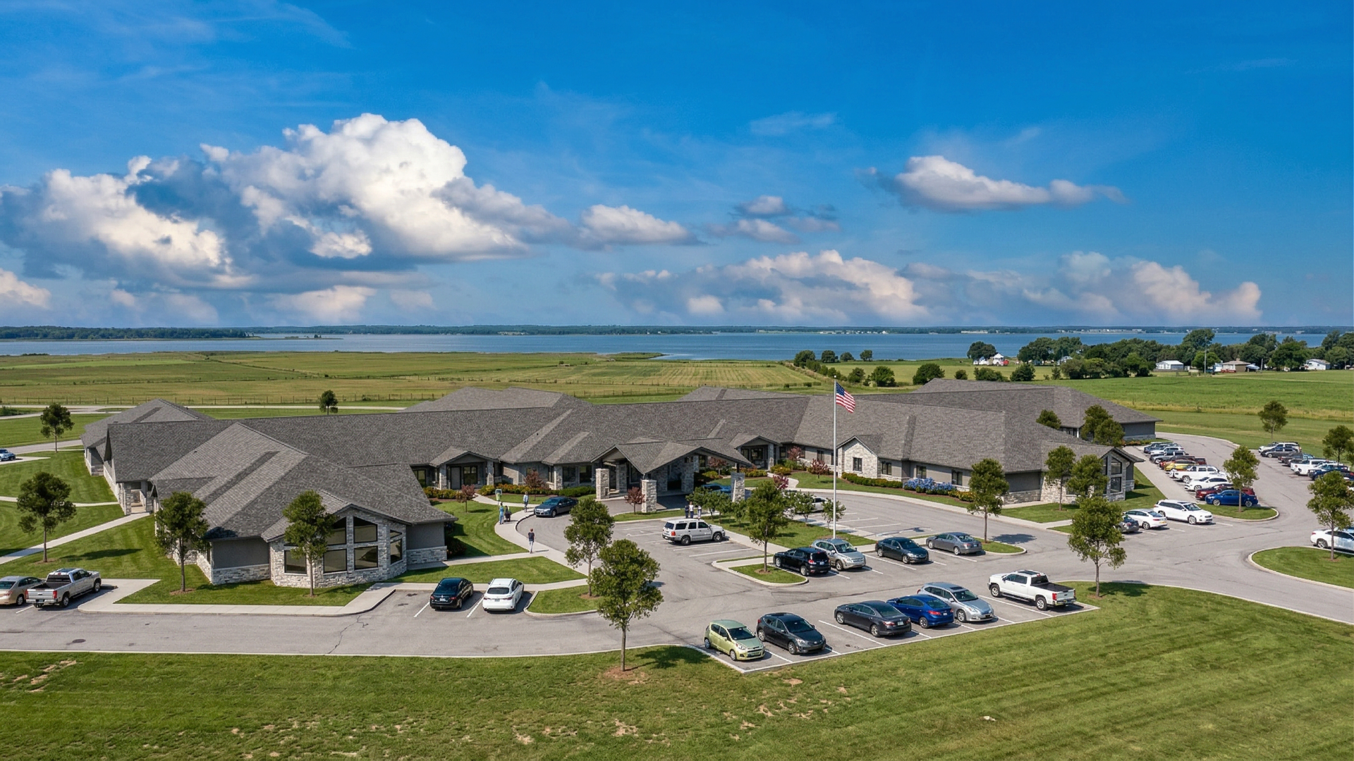 Aerial view of a senior care community complex with parked cars, green lawns, and a cloudy blue sky