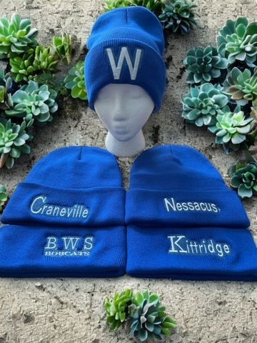 Four bright blue winter beanies arranged on a sandy background with succulents. Each features different white embroidery.