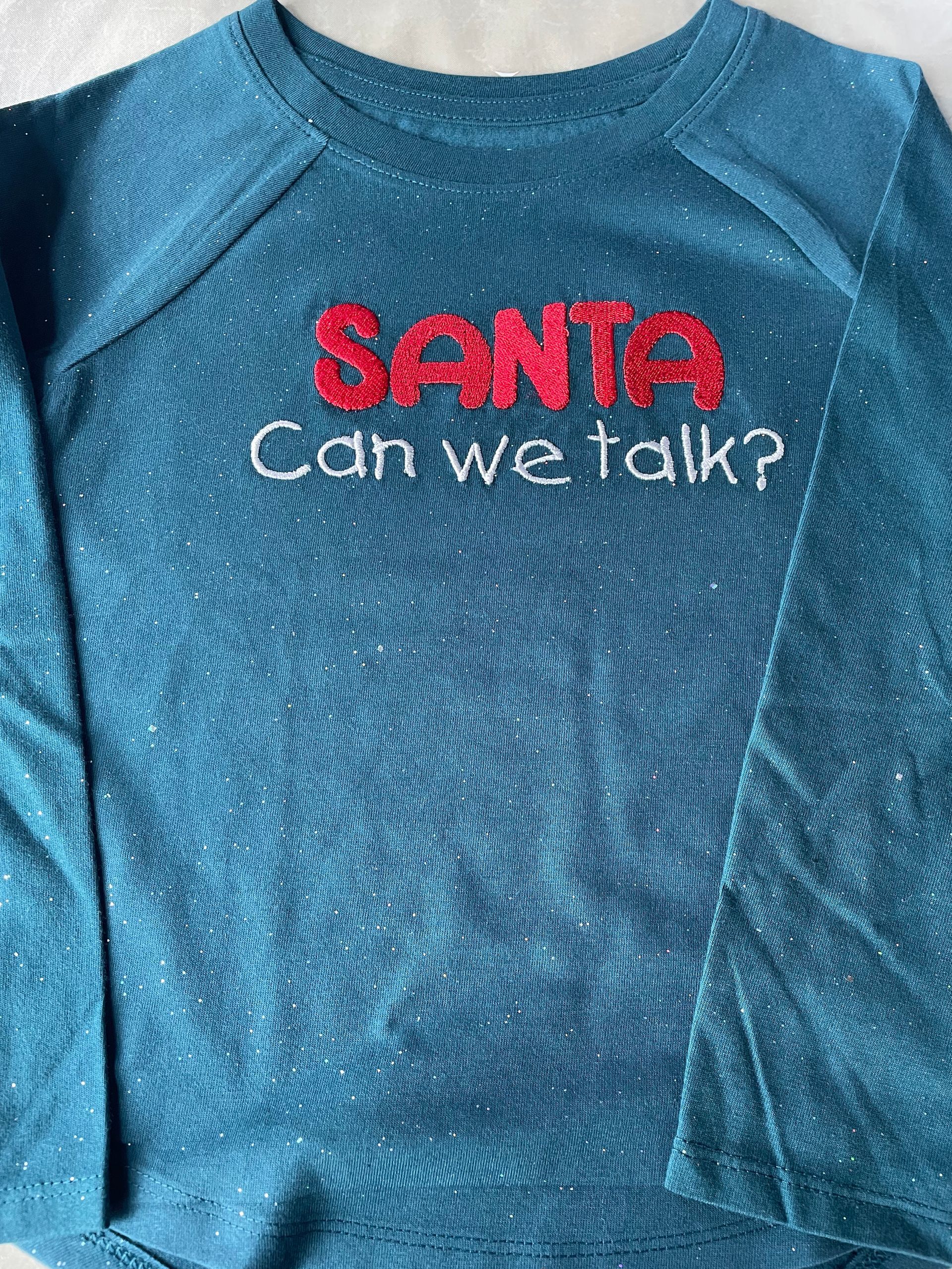 A teal, glittery long-sleeved shirt with the red sequined text 