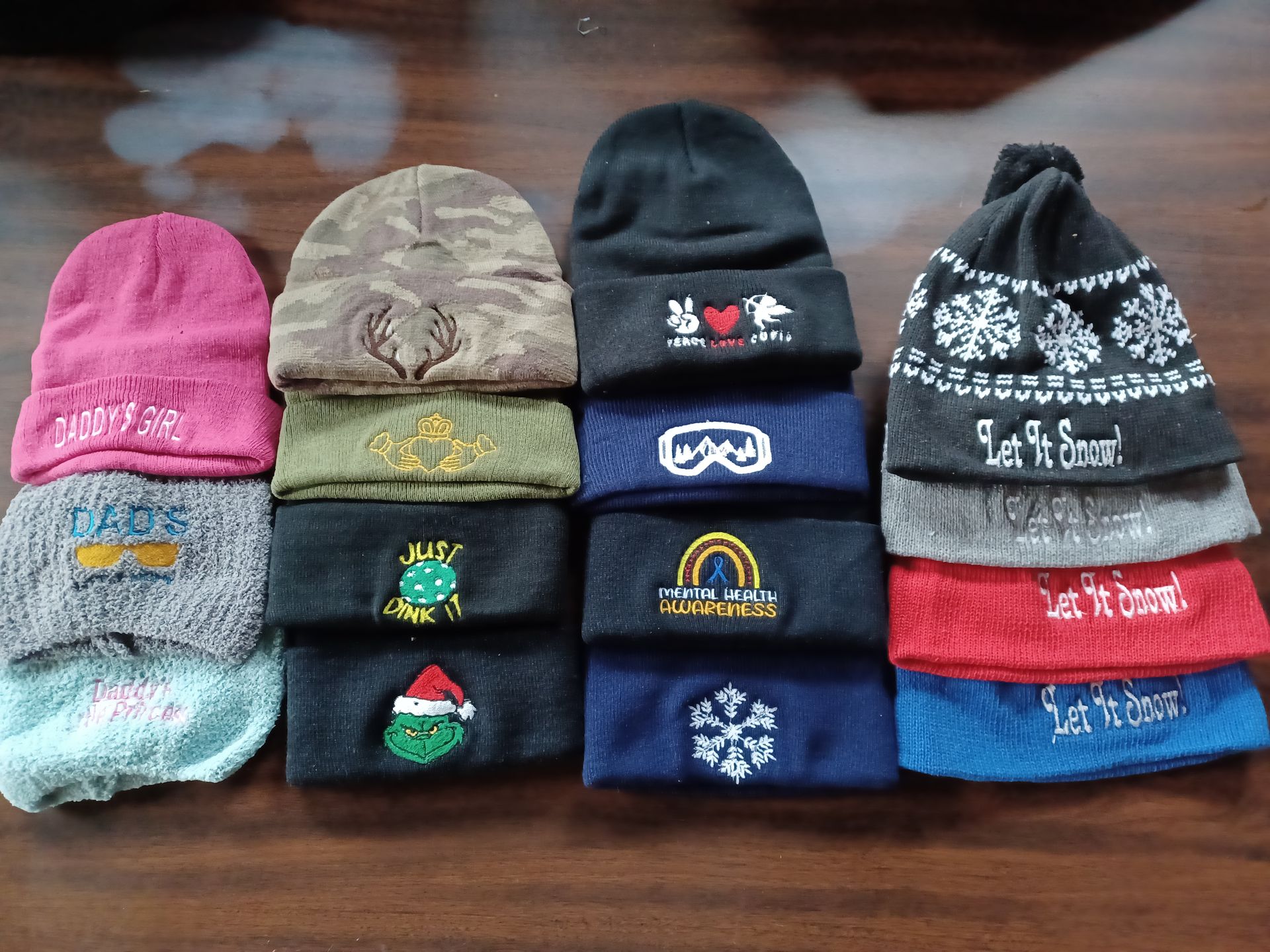 A collection of fourteen folded knit beanies in various colors and patterns arranged in four stacks on a wooden surface.