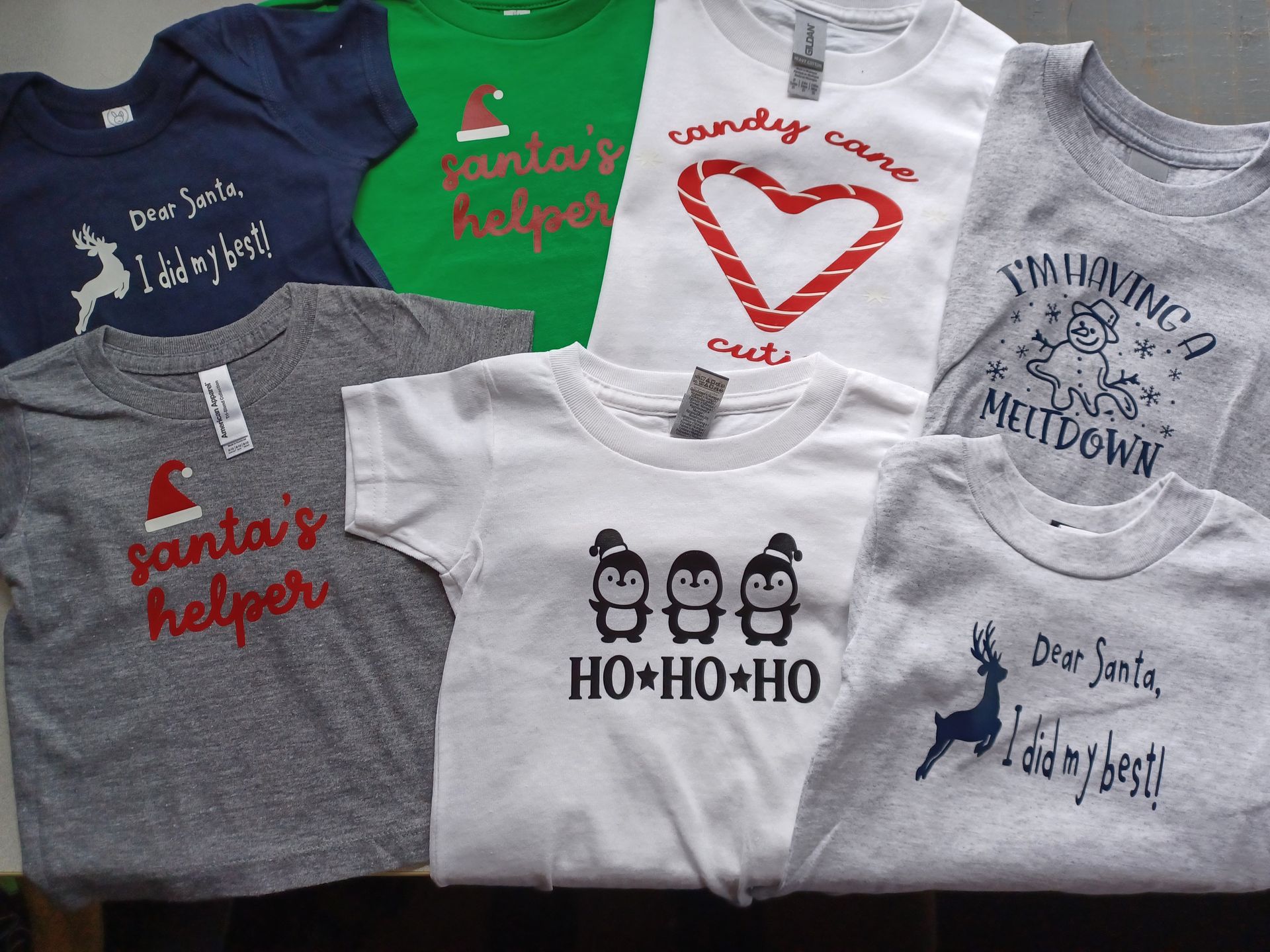 A flat lay of seven children's shirts with Christmas-themed designs in various colors and styles.