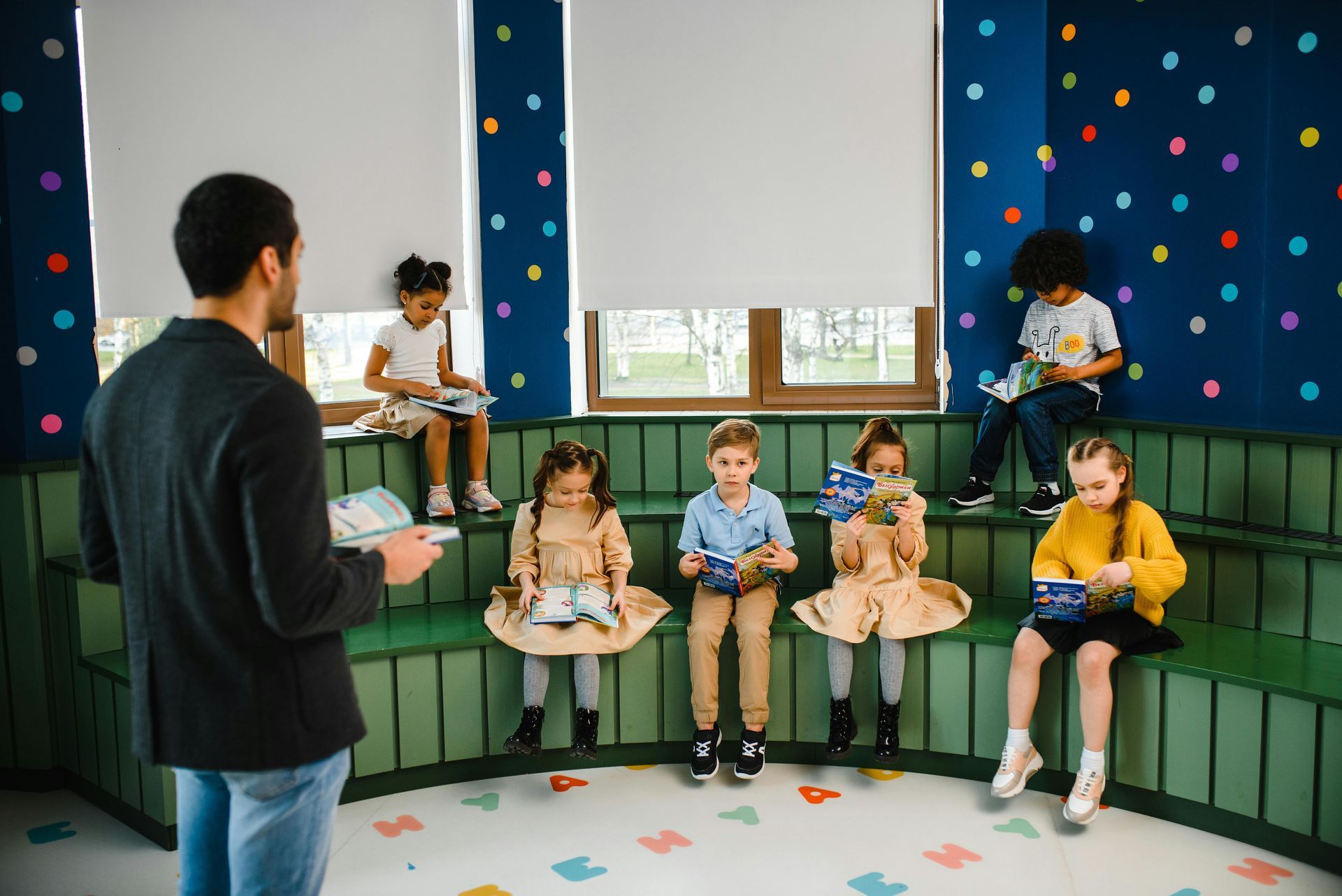 Children sitting in a room with polka dots with a teacher reading to them.