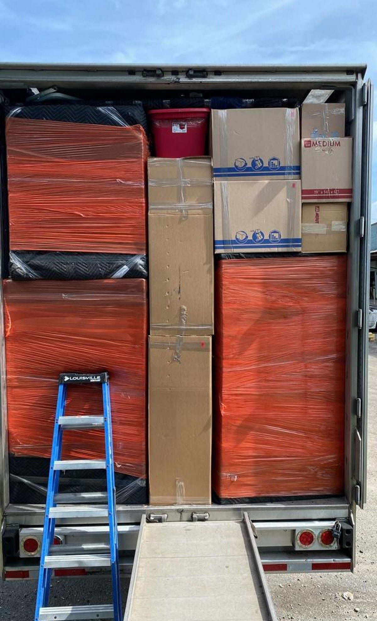 A truck cargo area filled with packed boxes and large orange-wrapped items, with a blue ladder in front.