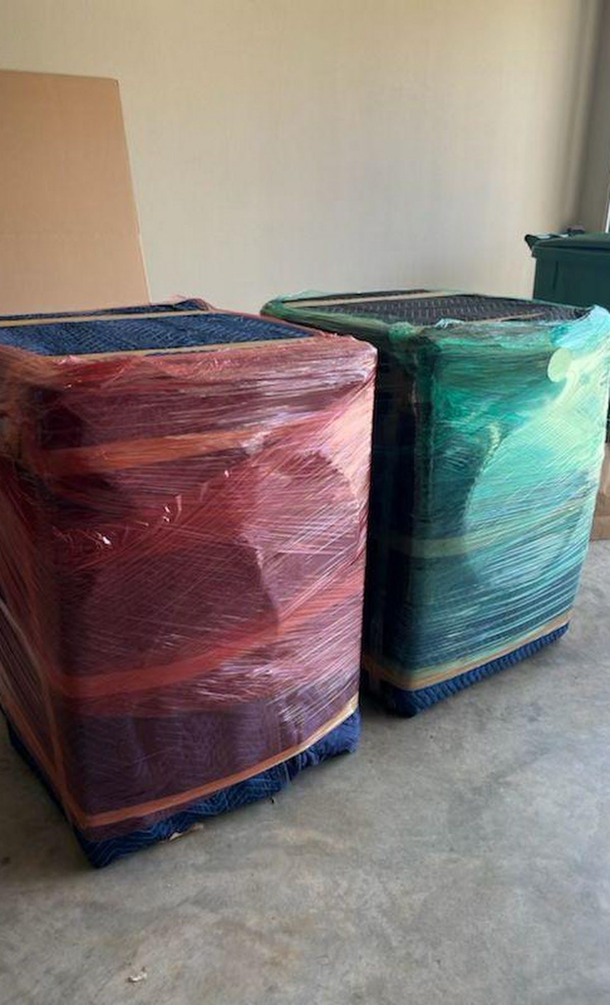 Two wrapped square ottomans, one red and one green, are ready for moving, sitting on a concrete floor.